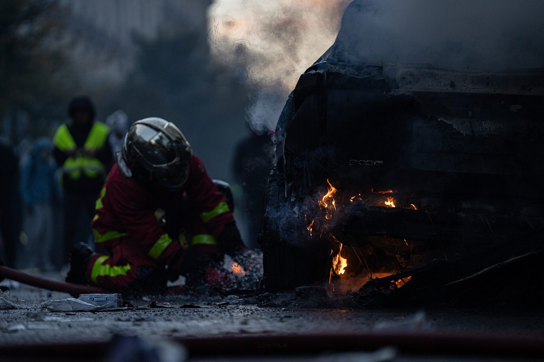 A firefighter in a helmet crouches beside a smouldering burnt-out car on a Paris street