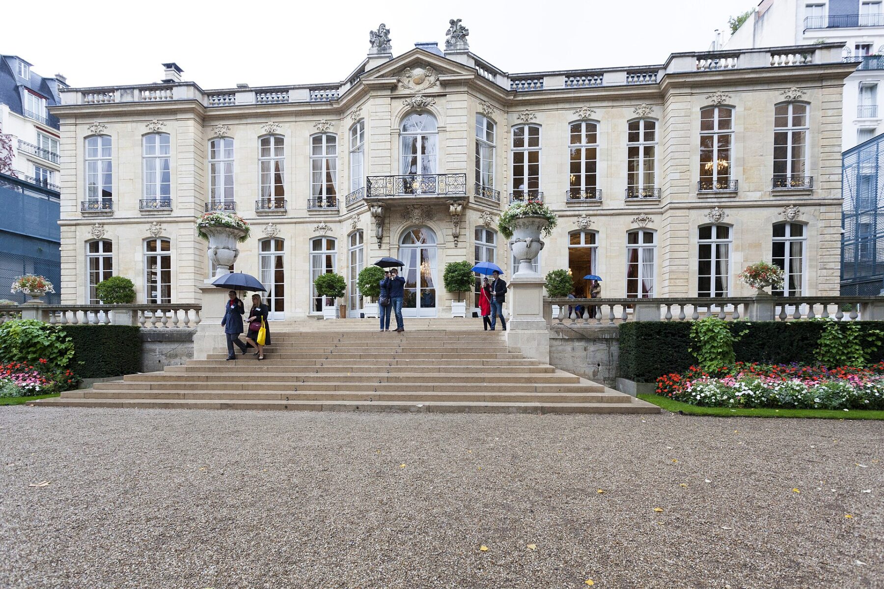 The stone facade and front steps of the Hôtel de Matignon in Paris on a rainy day, with visitors holding umbrellas