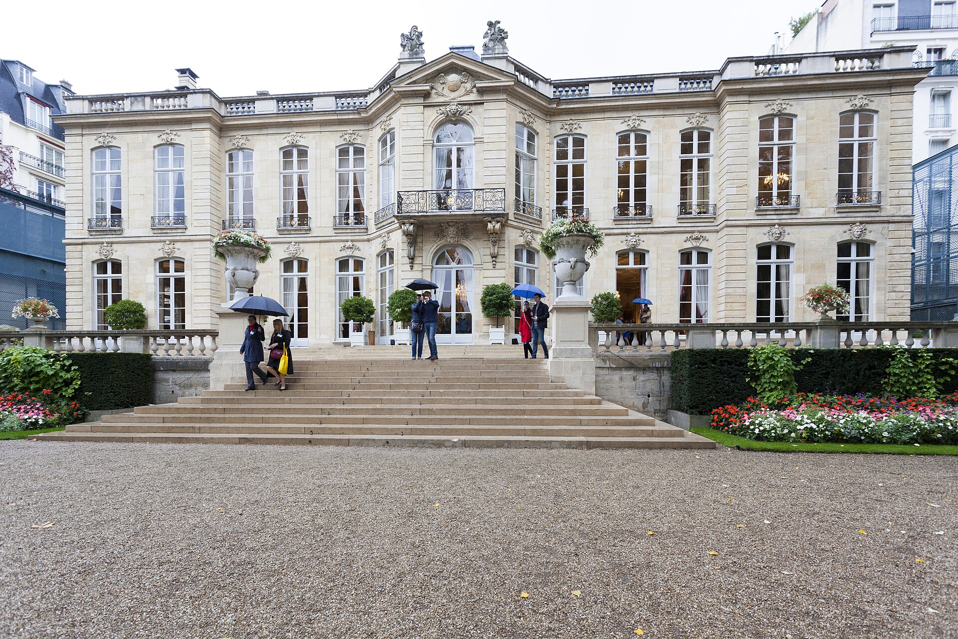 The stone facade and front steps of the Hôtel de Matignon in Paris on a rainy day, with visitors holding umbrellas