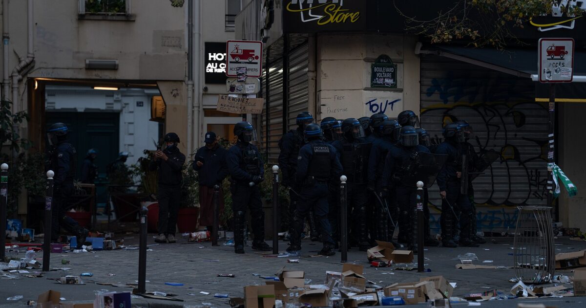 Riot police in helmets stand on a Paris street corner beside a looted shop with debris on the pavement
