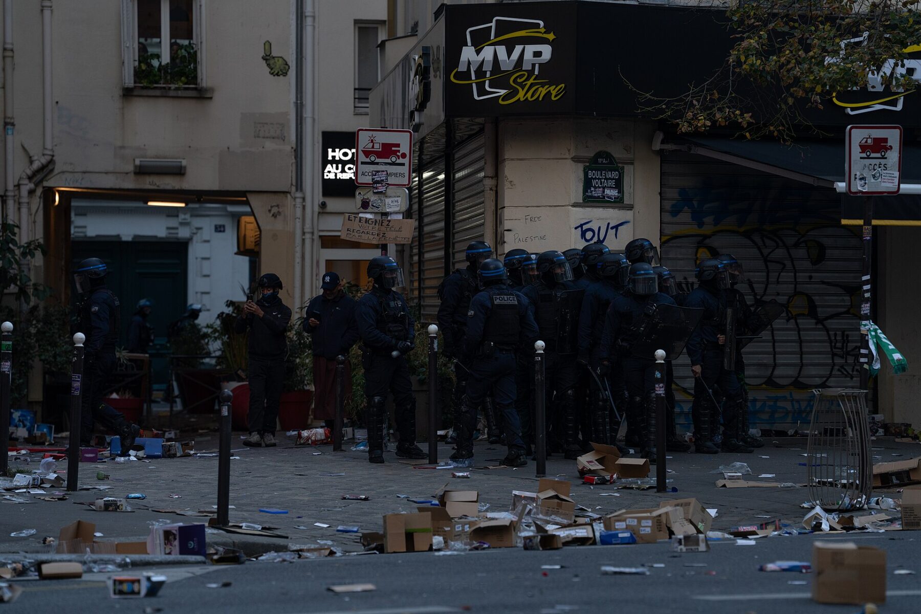 Riot police in helmets stand on a Paris street corner beside a looted shop with debris on the pavement