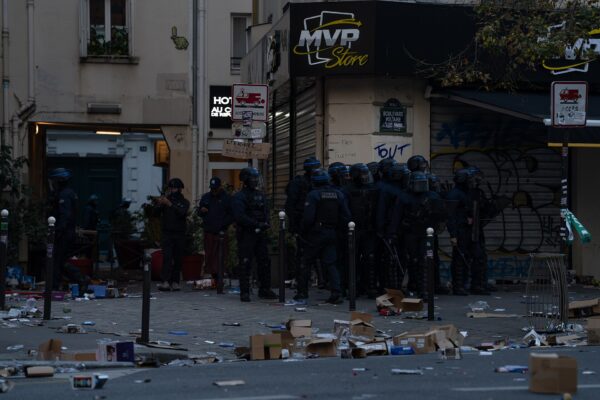 Riot police in helmets stand on a Paris street corner beside a looted shop with debris on the pavement