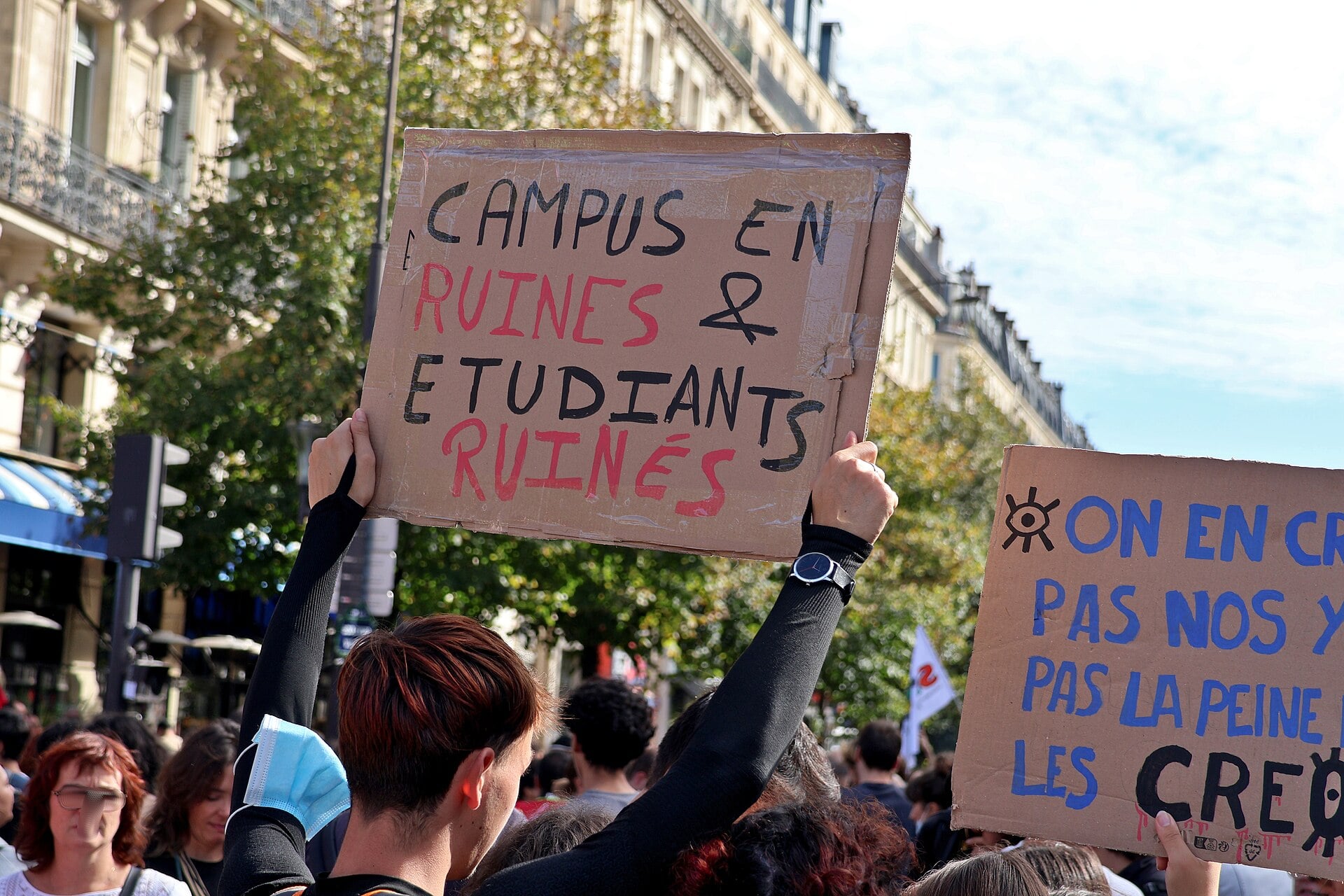 Young protesters in Paris hold up cardboard signs, one reading Campus en ruines et étudiants ruinés, under plane trees on a sunny day
