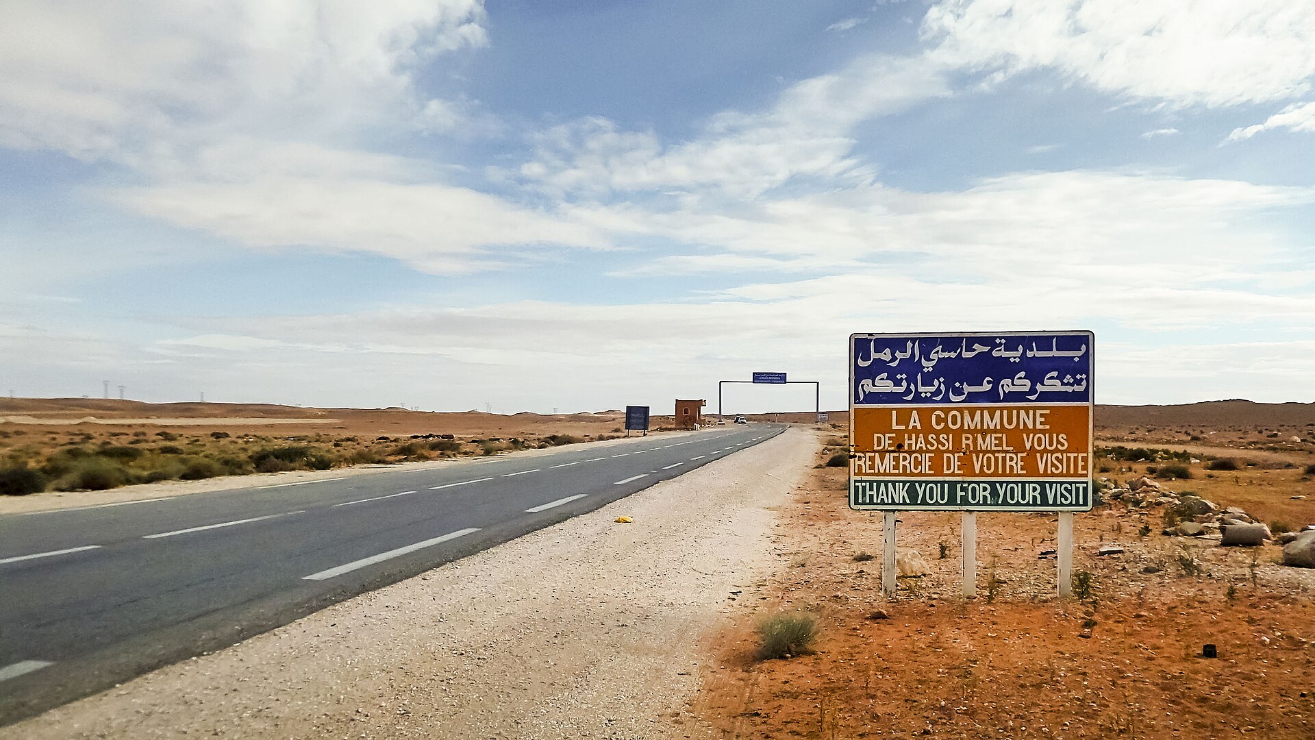 A road and a bilingual Arabic and French sign at the entrance of Hassi R'Mel in the Algerian desert