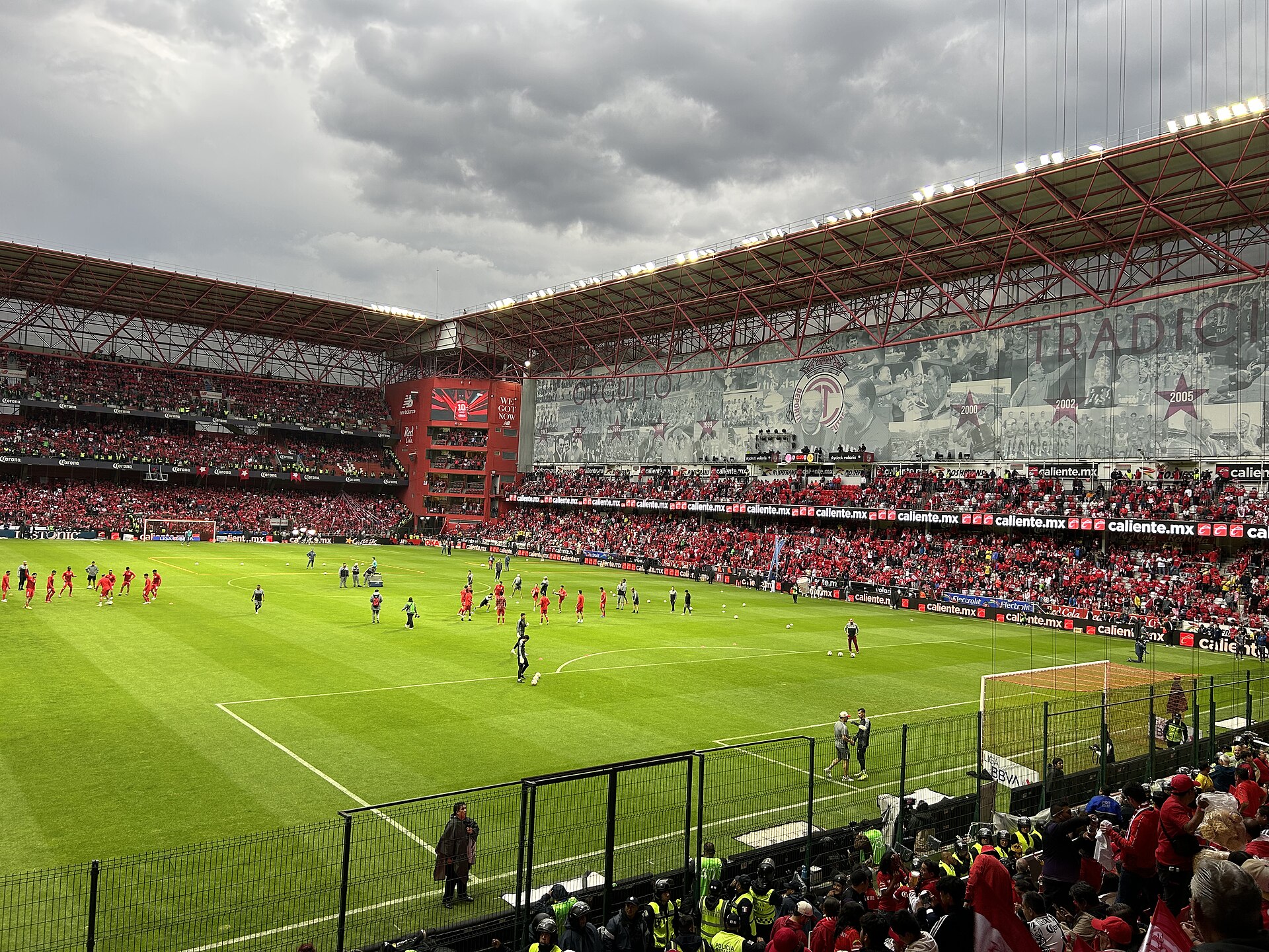 Players warming up on the pitch at Estadio Nemesio Díez in Toluca, with full red-clad stands under a red steel roof and a large mural on the main stand