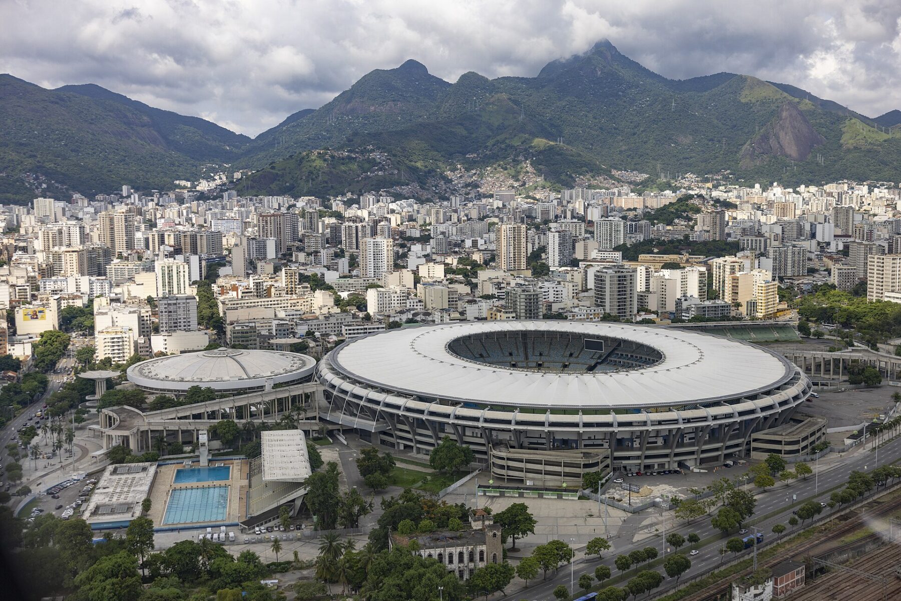 Aerial view of the white-roofed Maracanã stadium in Rio de Janeiro, with apartment blocks and green mountains behind under a cloudy sky