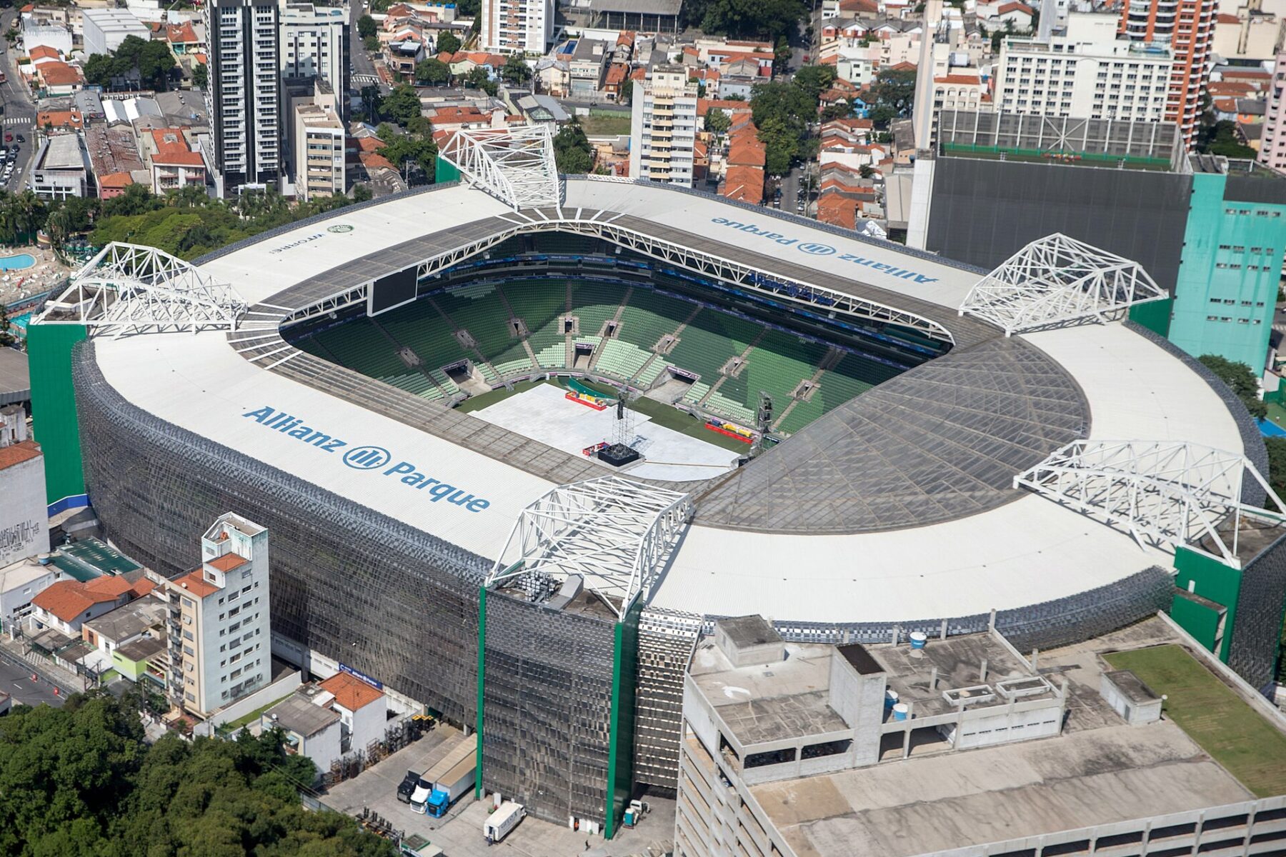 Aerial view of a rectangular stadium with a white roof, steel lattice supports and green seats, its pitch covered for an event, surrounded by city buildings