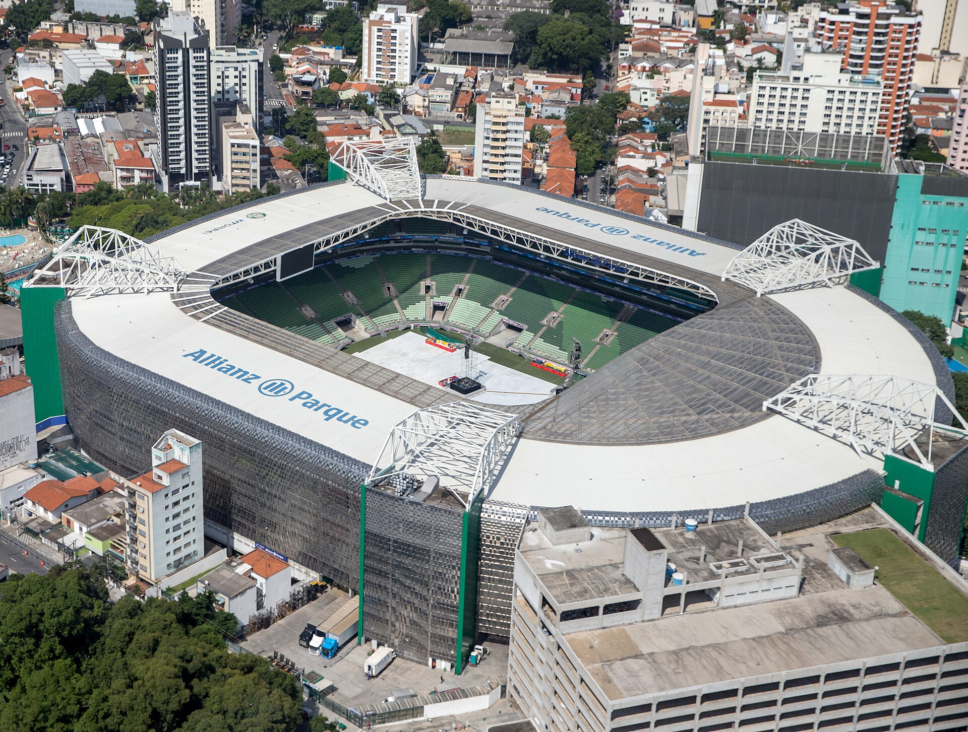 Aerial view of a rectangular stadium with a white roof, steel lattice supports and green seats, its pitch covered for an event, surrounded by city buildings