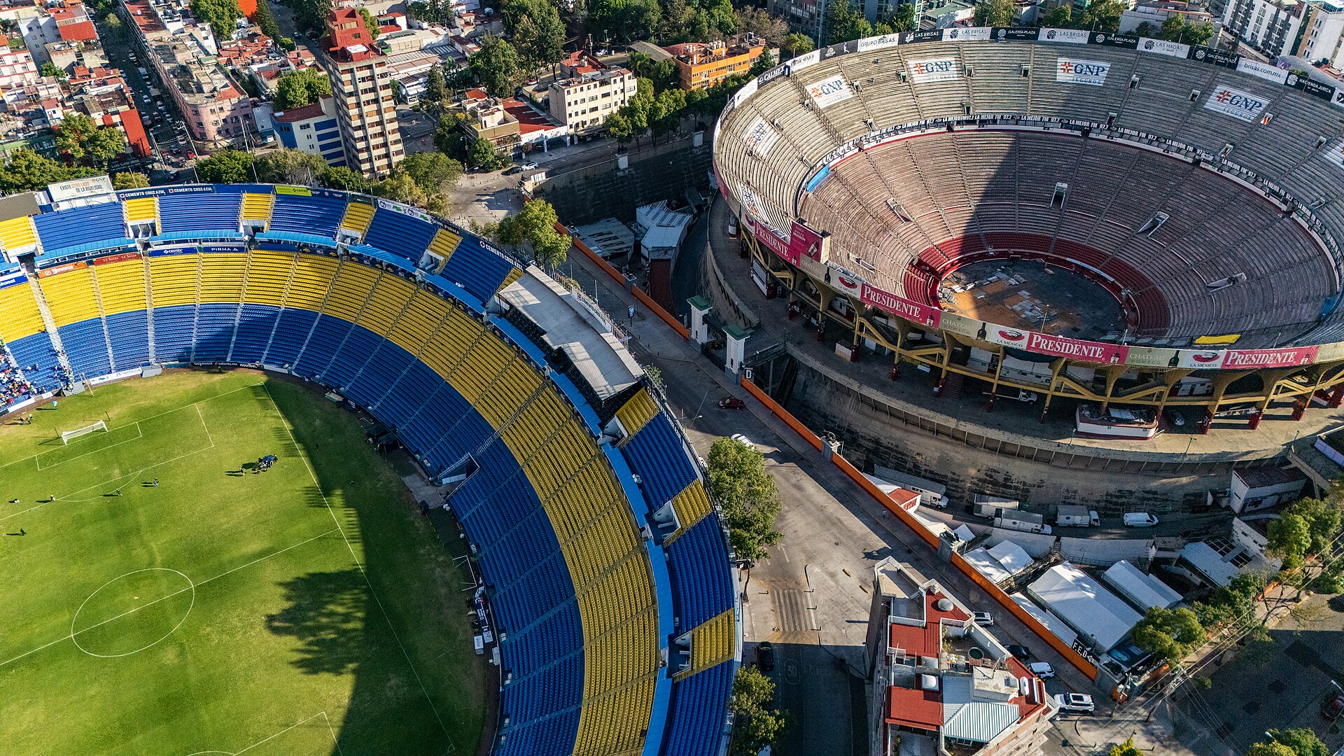 Aerial view of the blue and yellow stands of the Estadio Ciudad de los Deportes on the left and the round Plaza México bullring on the right, separated by a street