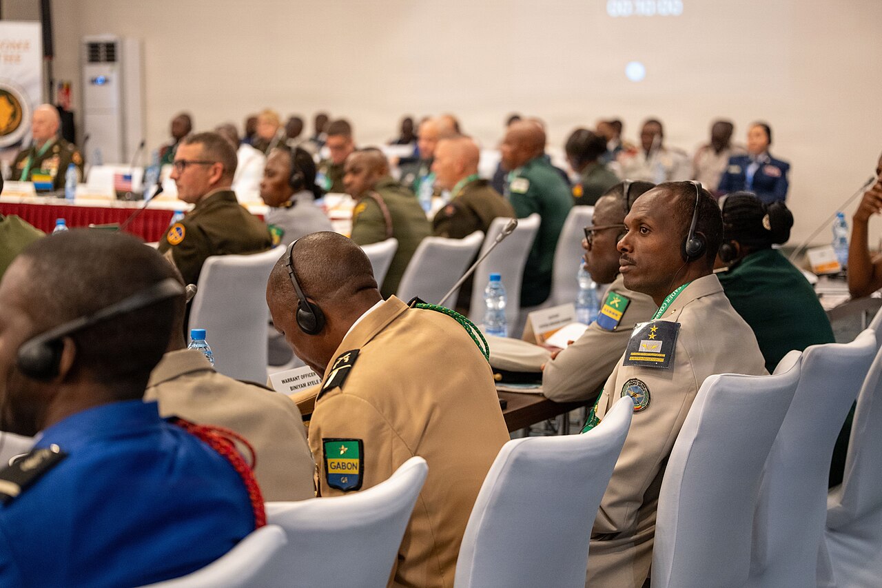 Gabonese and other military officers in uniform and headsets listen at a conference table in Libreville, with a Gabon flag patch on one sleeve