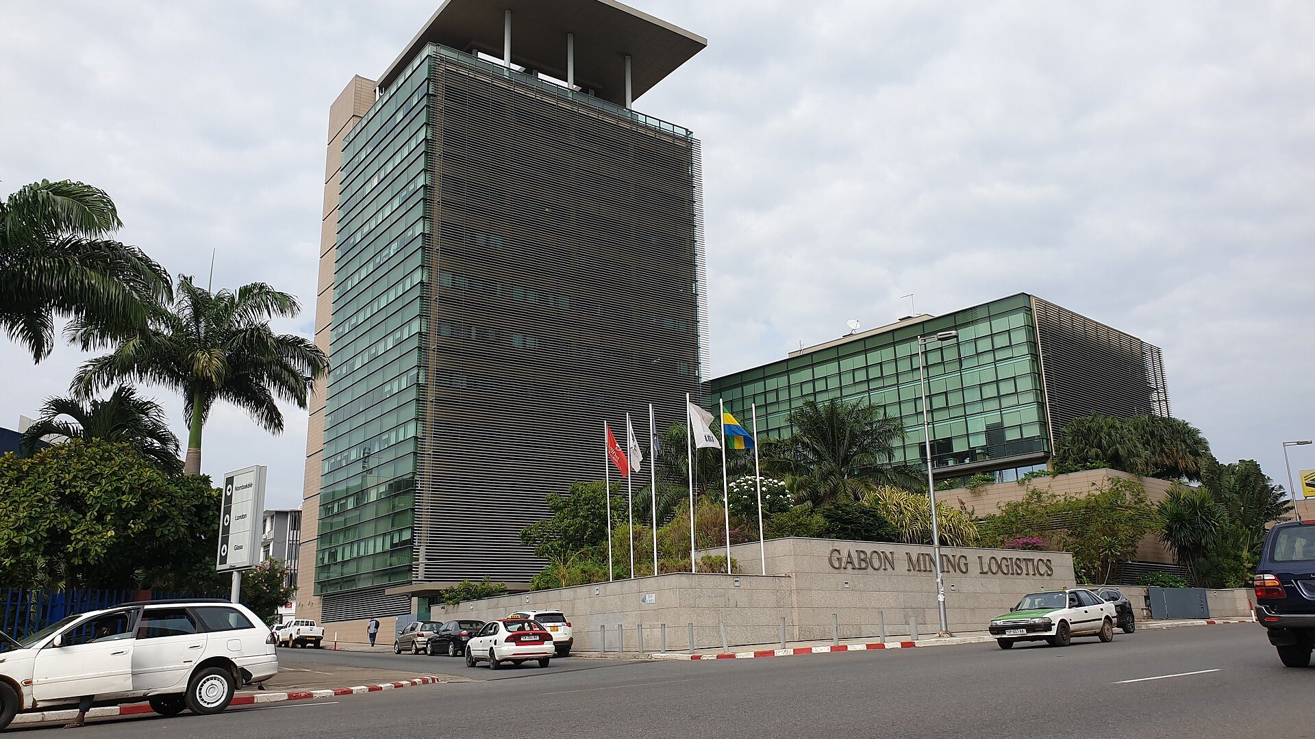 The glass Gabon Mining Logistics office tower and its sign beside a street in central Libreville, with the Gabonese flag and passing cars