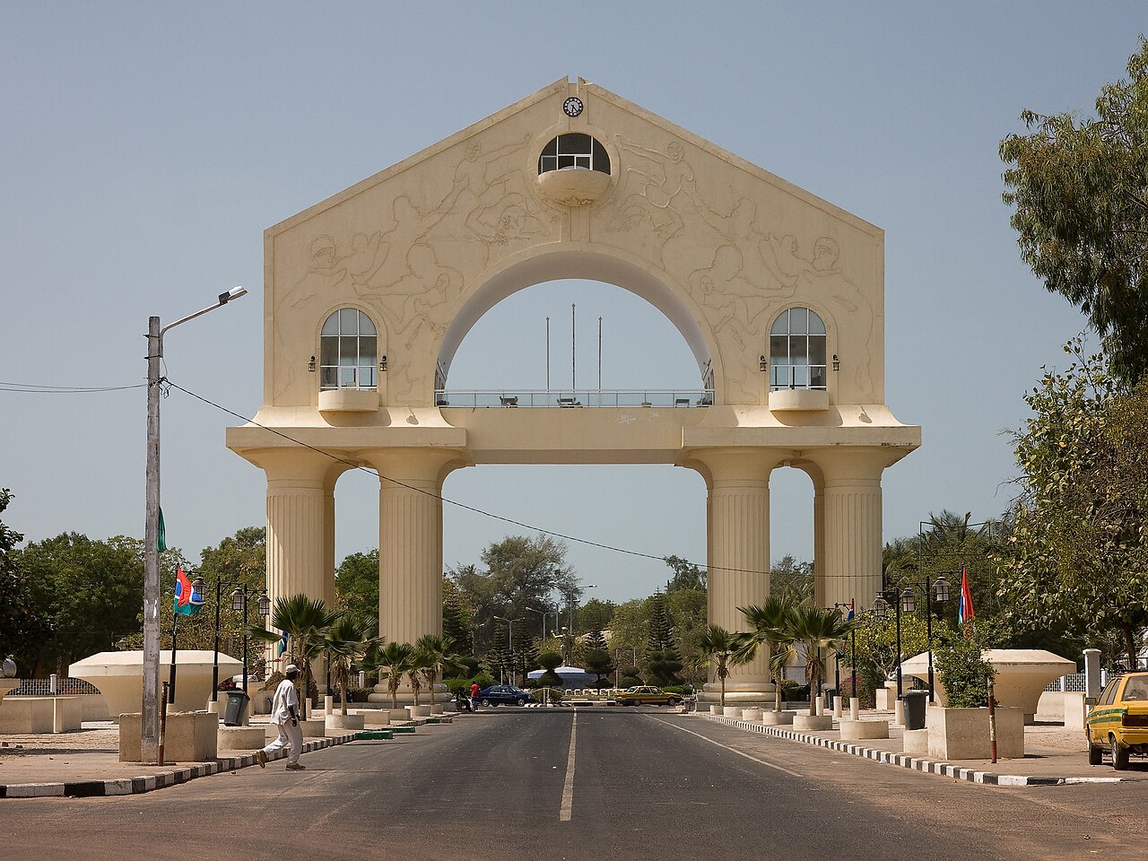 Arch 22, a large cream-coloured memorial arch on a road in Banjul, with a pedestrian and taxis beneath it