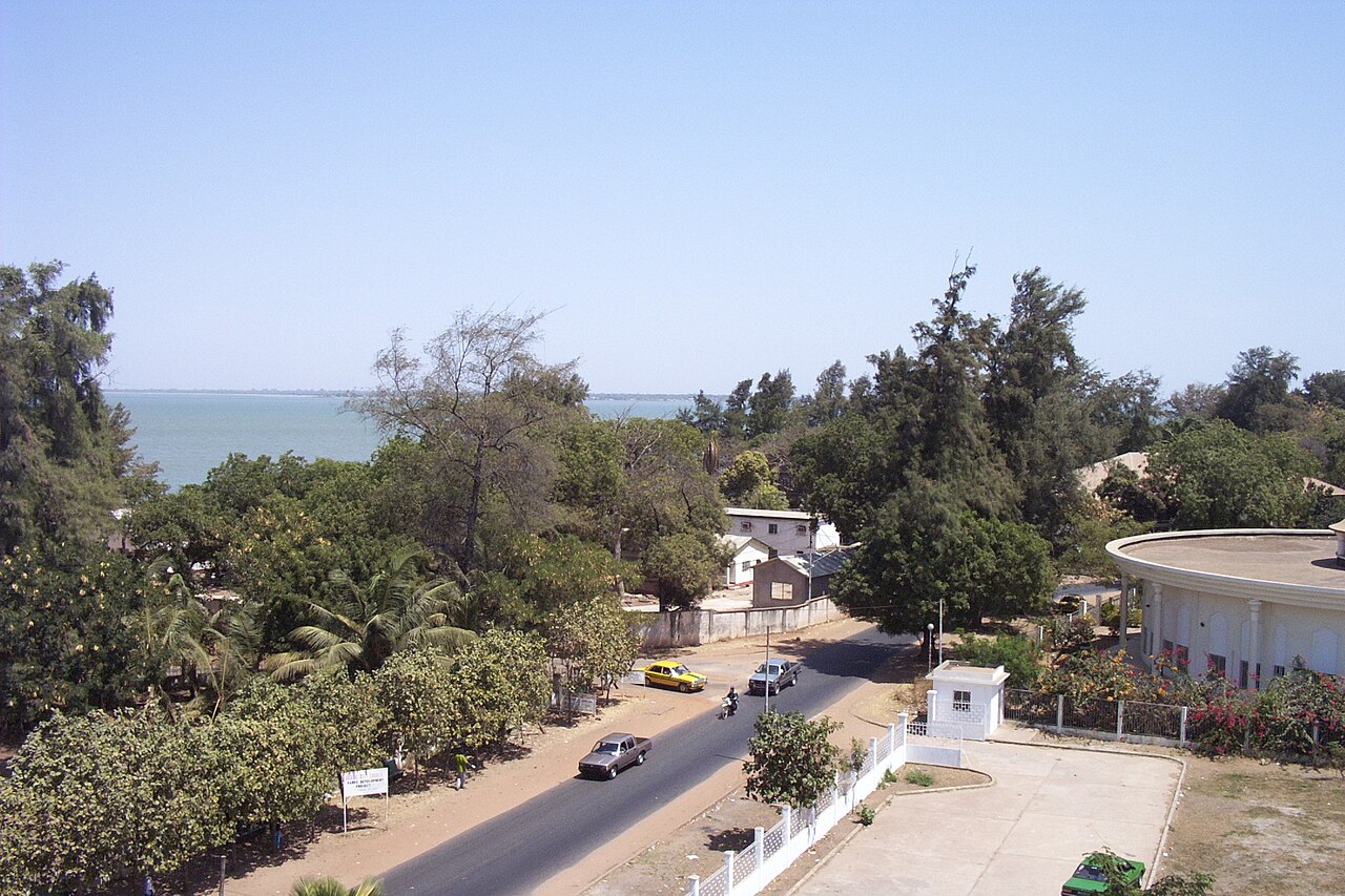 A tree-lined road in Banjul beside the Gambia River, with a taxi and cars on the street and a round building on the right.