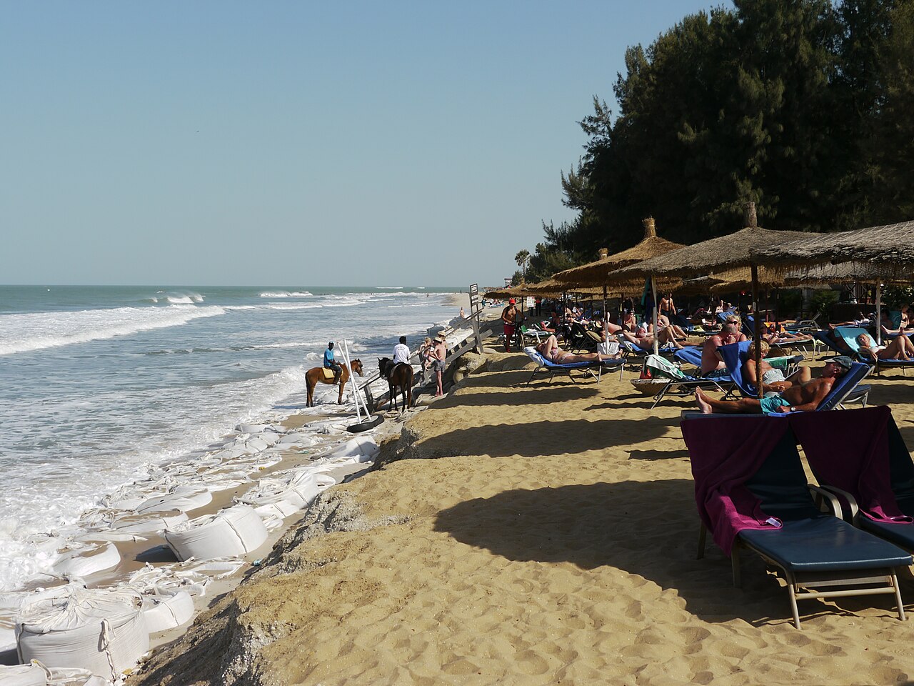 Sunbathers under thatched parasols on Kololi beach on The Gambia's Atlantic coast, with two horse riders at the waterline and sandbags against the surf.