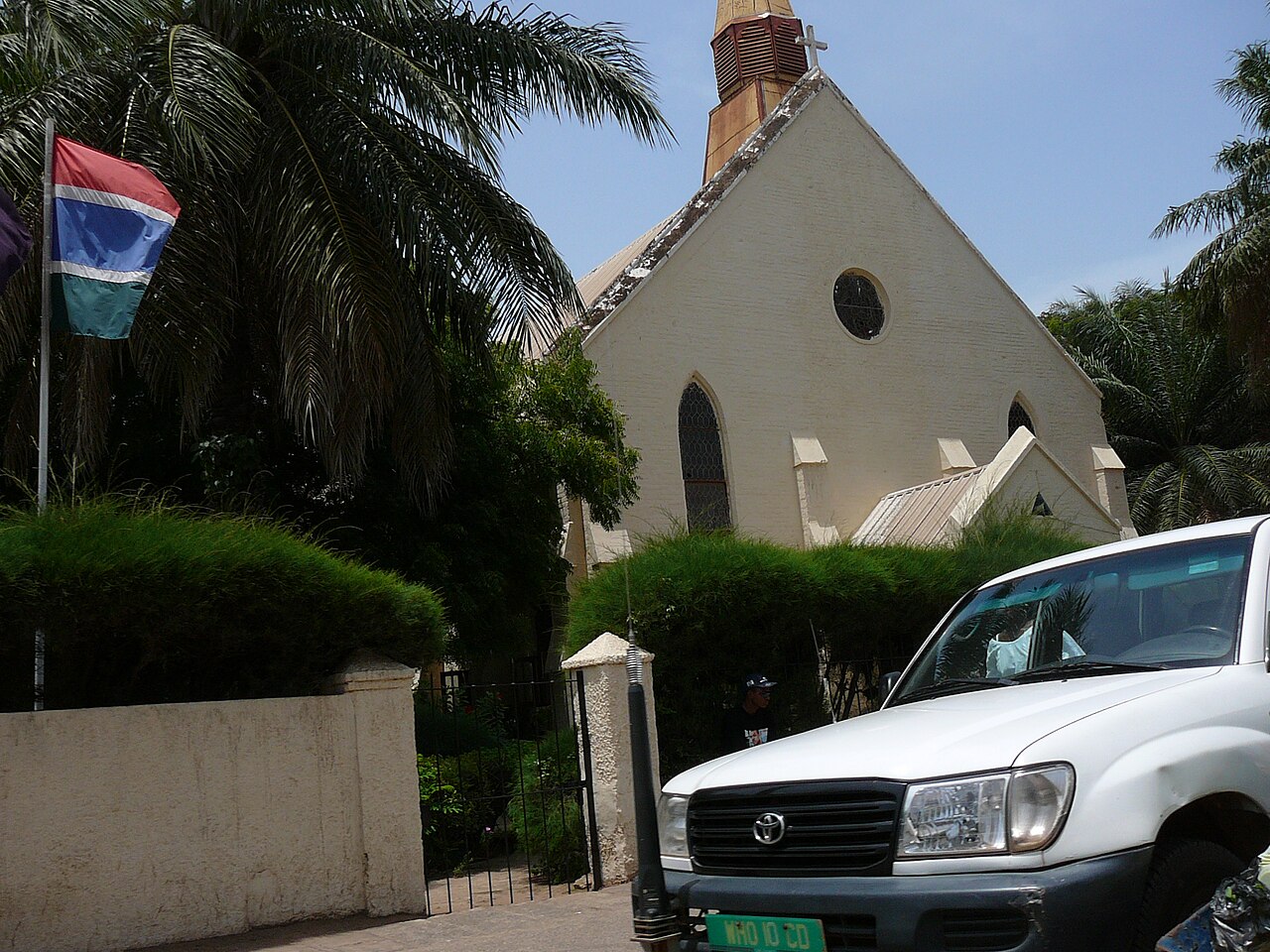 Gambian flag beside a cream-coloured church among palm trees in Banjul