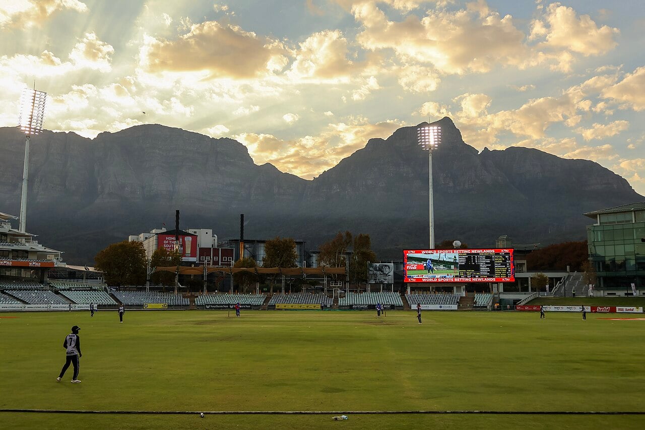 Newlands Cricket Ground in Cape Town in evening light, with players on the field, floodlights and the mountains of Cape Town behind the stands