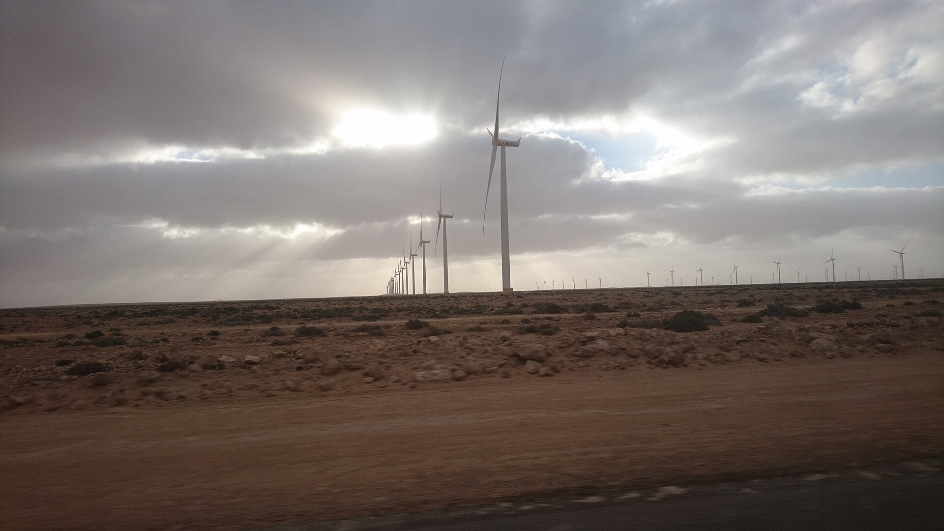 A row of wind turbines on flat stony desert near Tarfaya, Morocco, under a heavy cloudy sky with sun rays
