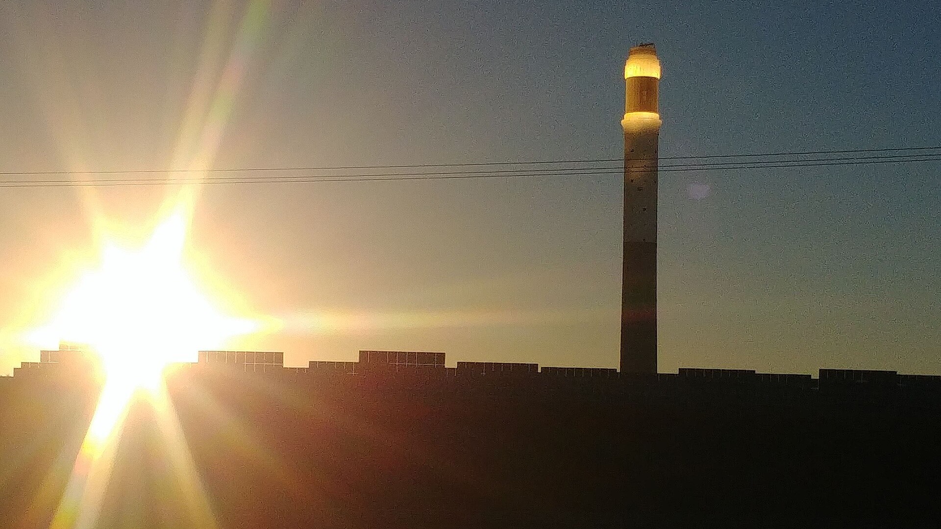 The Noor III solar tower lit at the top, with the setting sun and solar panels in the foreground