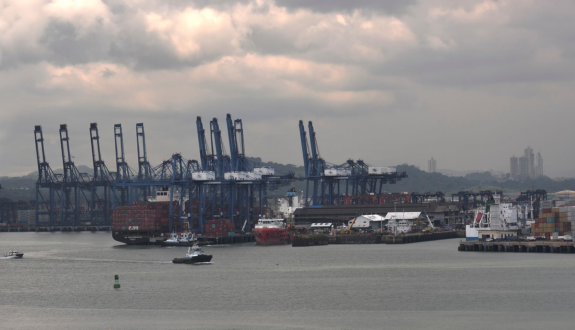 Blue container cranes at the Port of Balboa in Panama under a cloudy sky, with ships, tugboats and city buildings in the distance