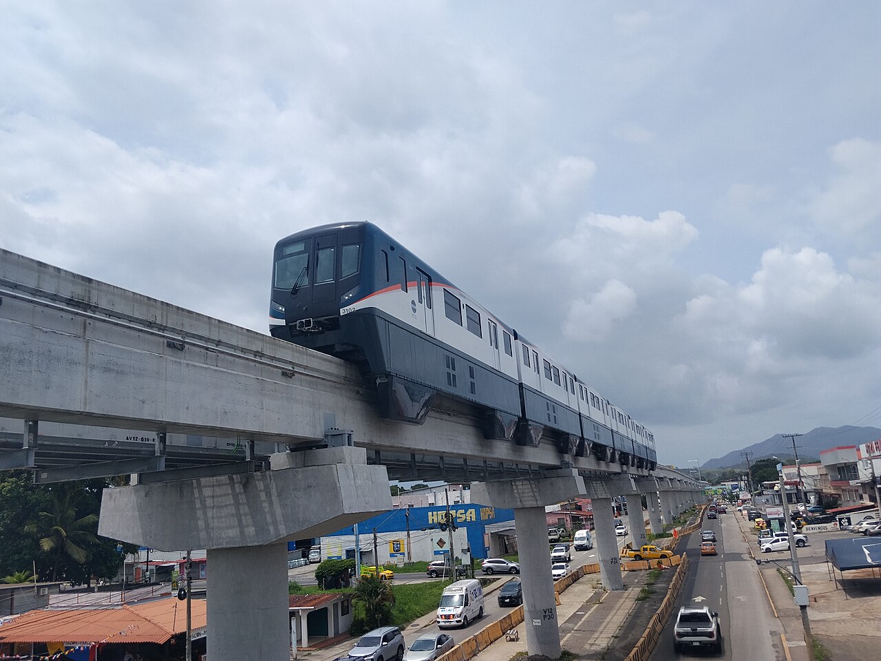 A dark blue and white monorail train on an elevated concrete track above a road with cars, under a cloudy sky