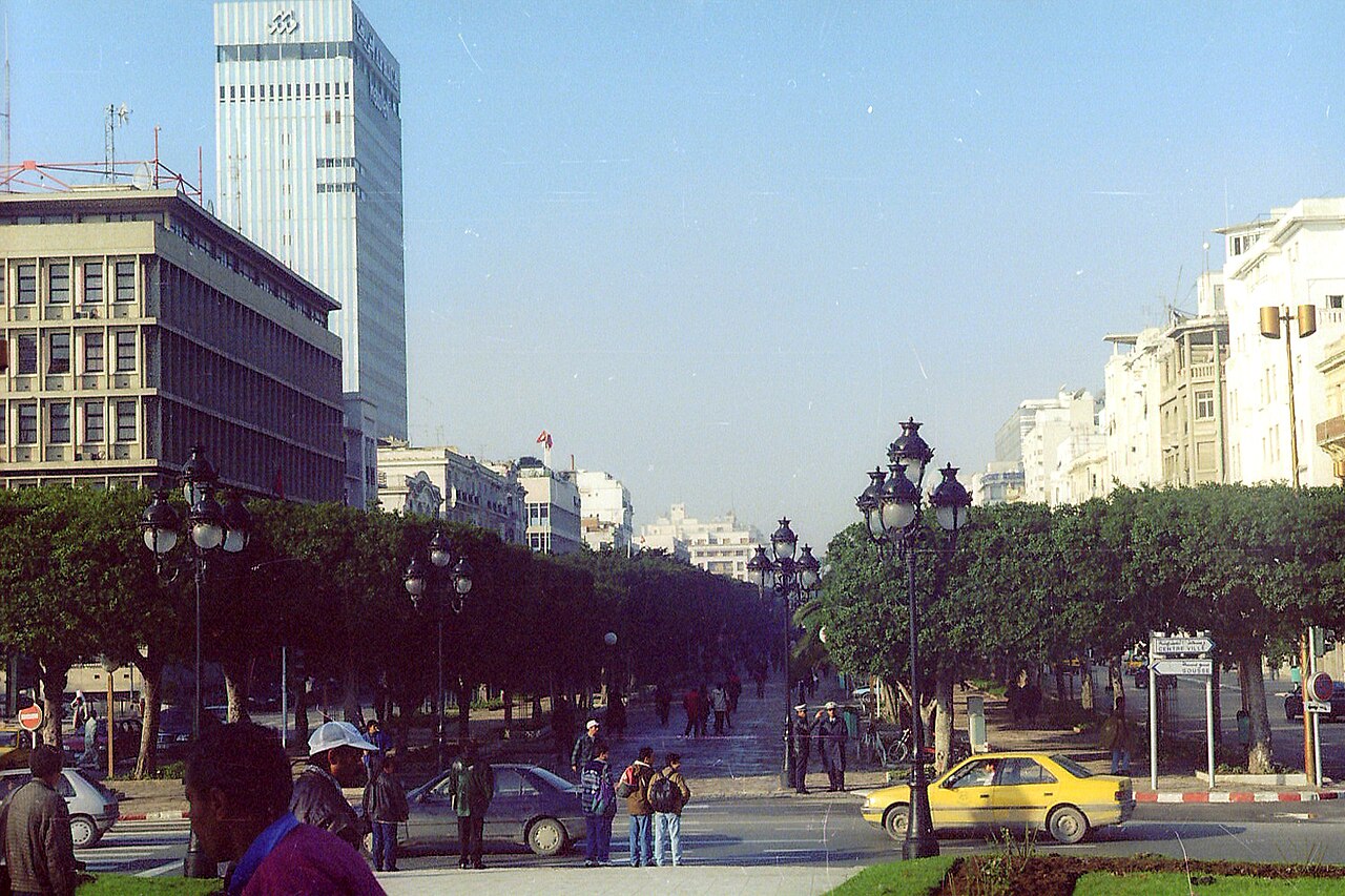 A wide tree-lined boulevard in central Tunis with pedestrians, a yellow taxi and office towers