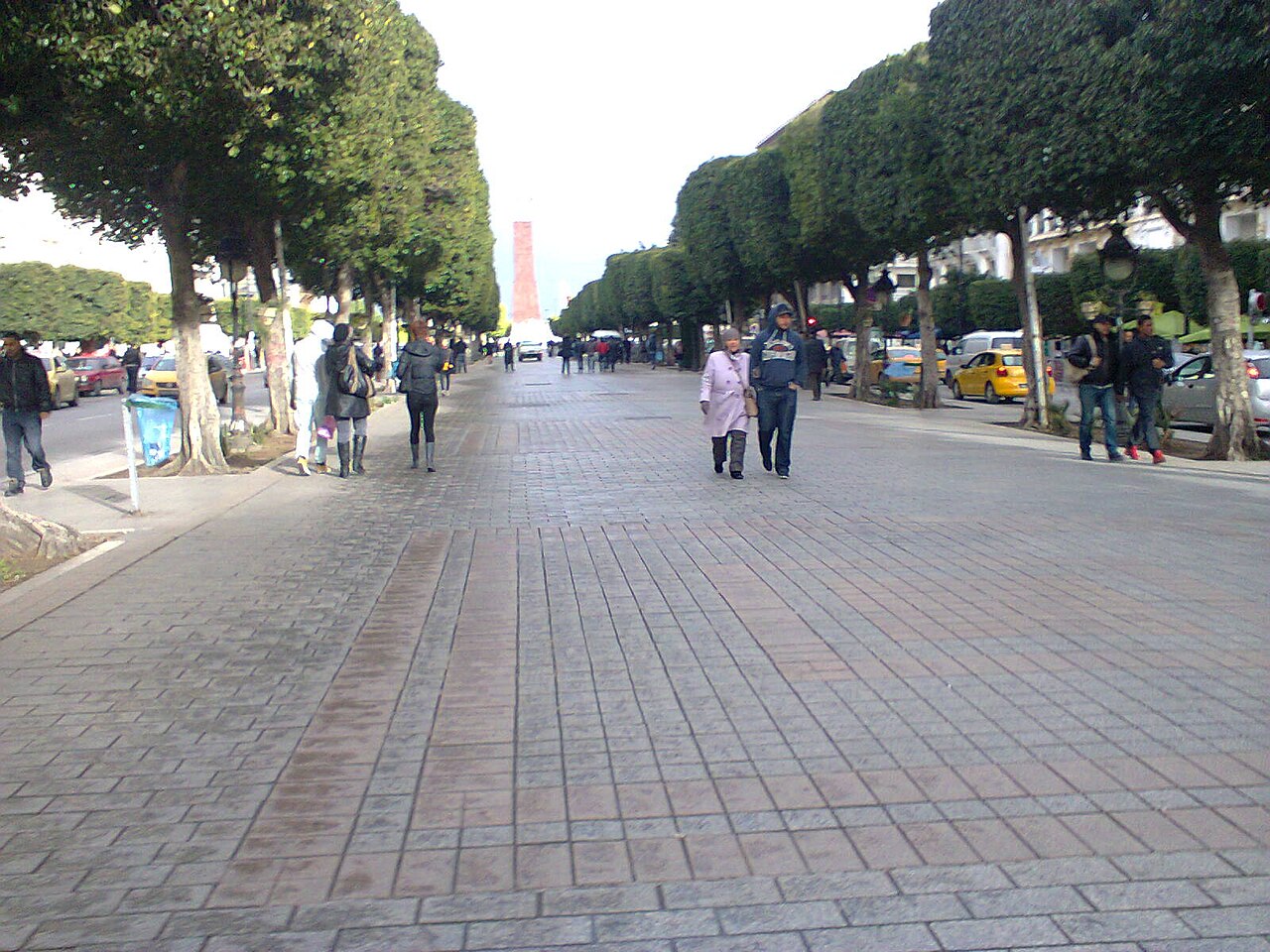 Pedestrians on a wide paved boulevard lined with trimmed trees in central Tunis, with a tall tower in the distance