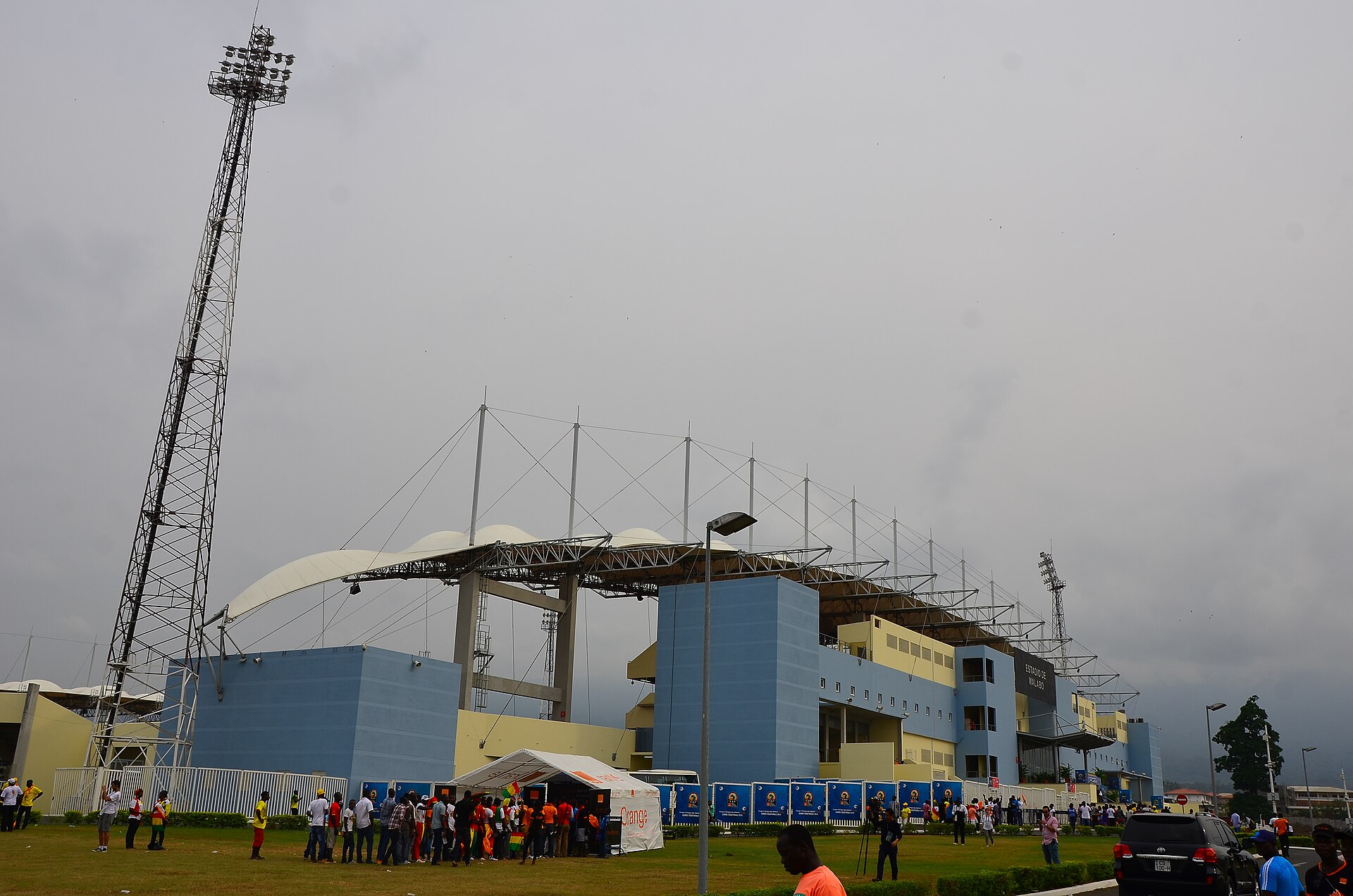 The new stadium in Malabo with a floodlight mast, a white roof canopy and spectators gathered on the grass in front under a grey sky