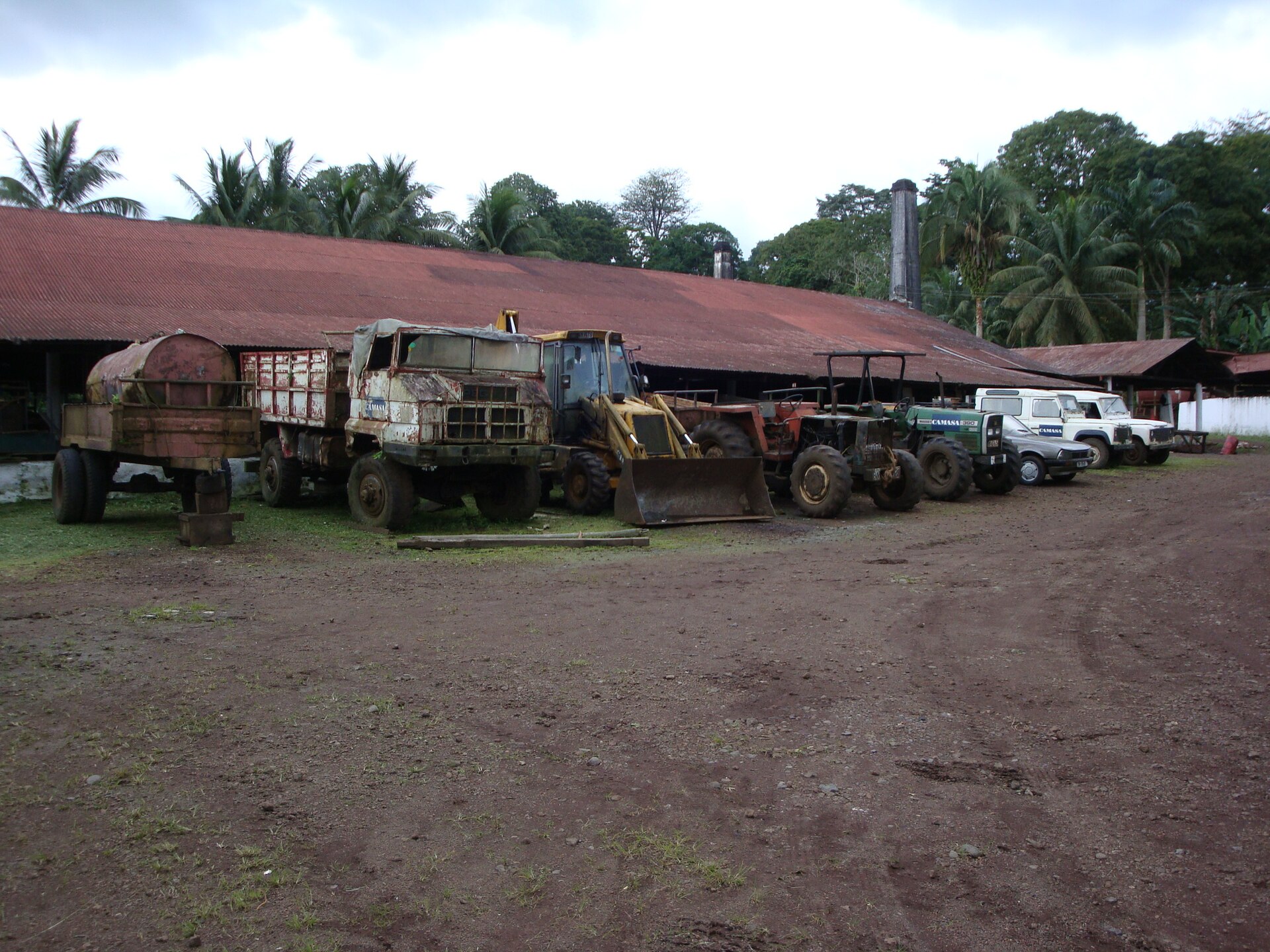 Trucks, a loader and tractors parked in front of a long rust-red roofed building surrounded by palm trees