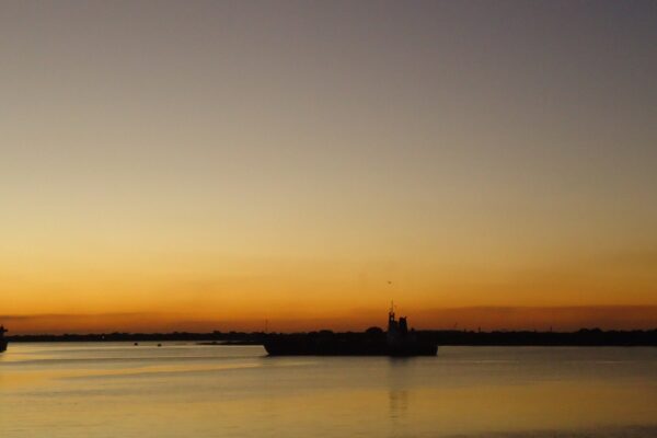 A cargo ship silhouetted on the bay of Asunción under an orange sky, with a second vessel at the left edge