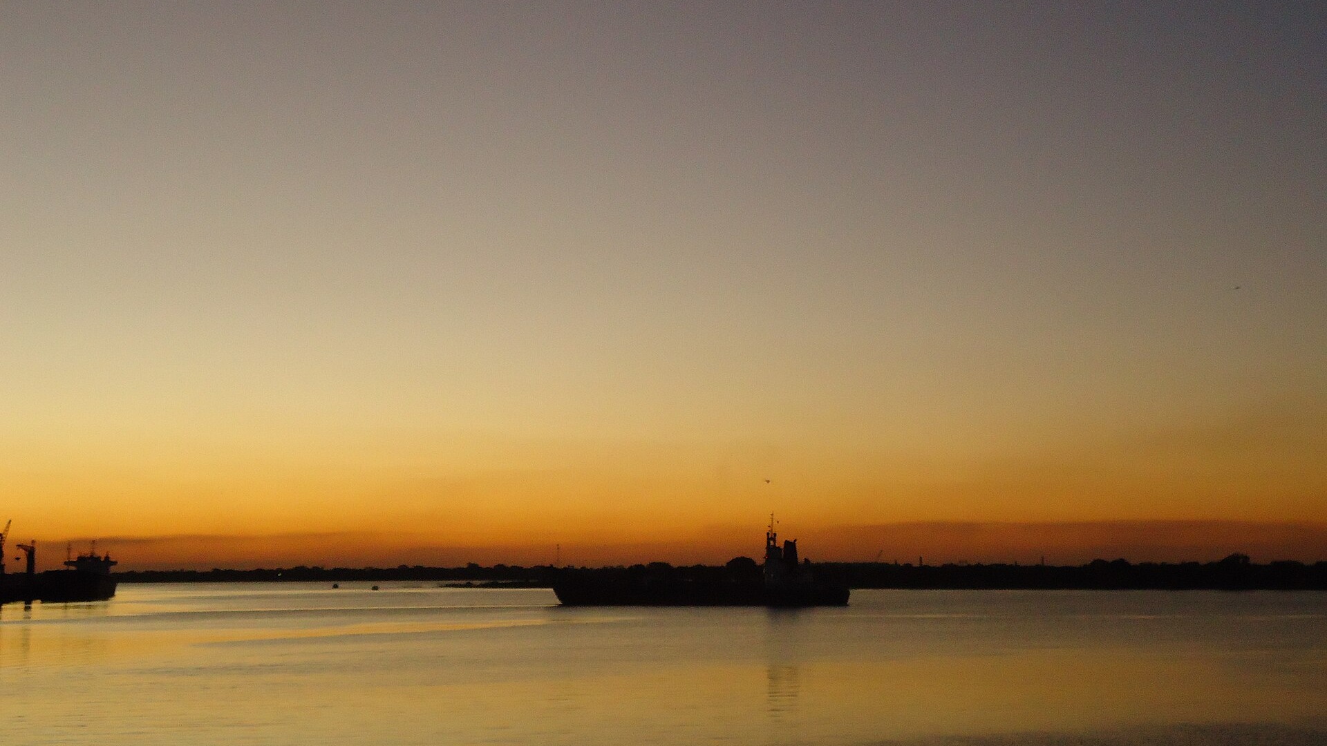 A cargo ship silhouetted on the bay of Asunción under an orange sky, with a second vessel at the left edge