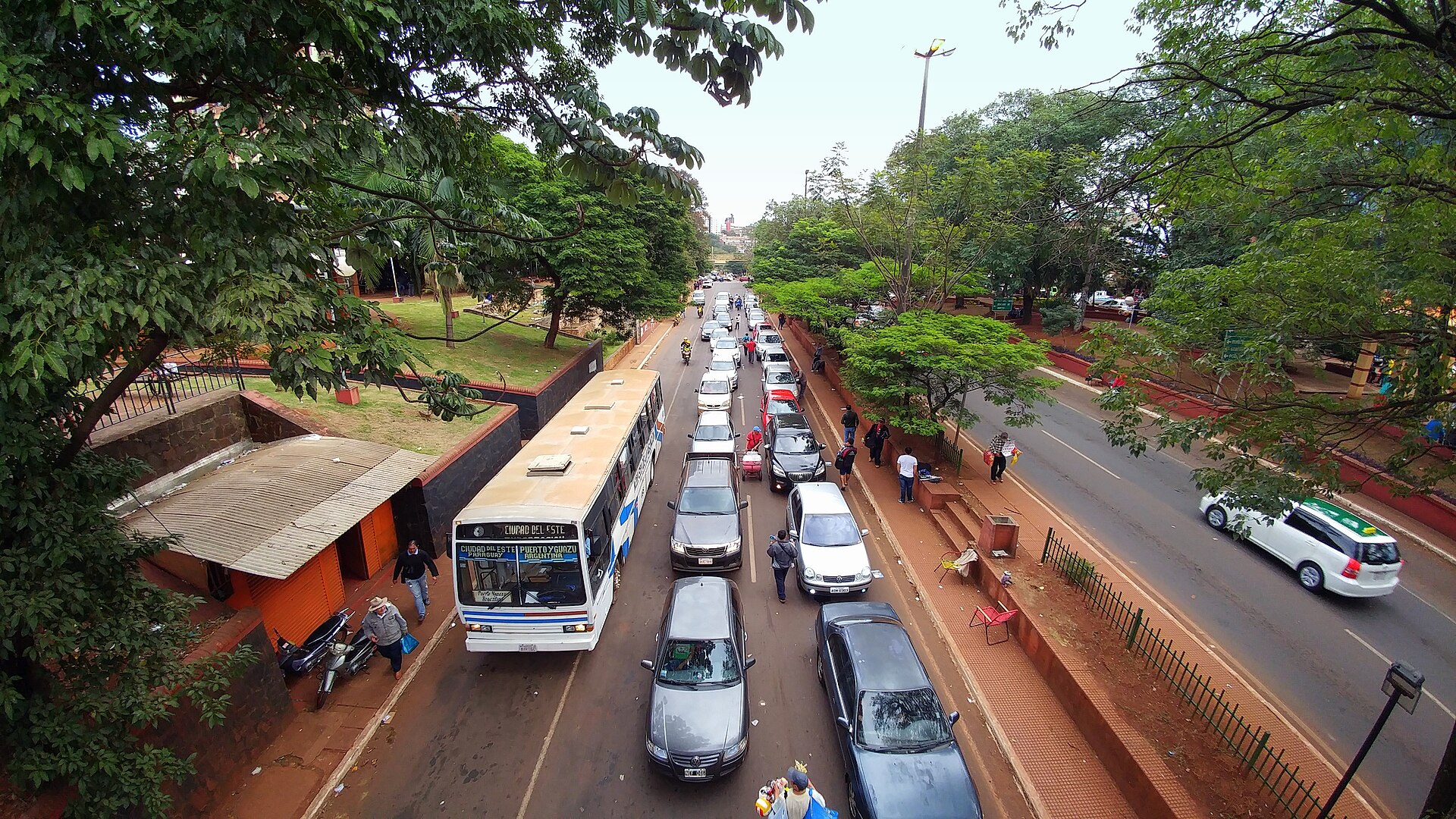 Cars, a city bus and pedestrians on a tree-lined road in Ciudad del Este seen from a footbridge