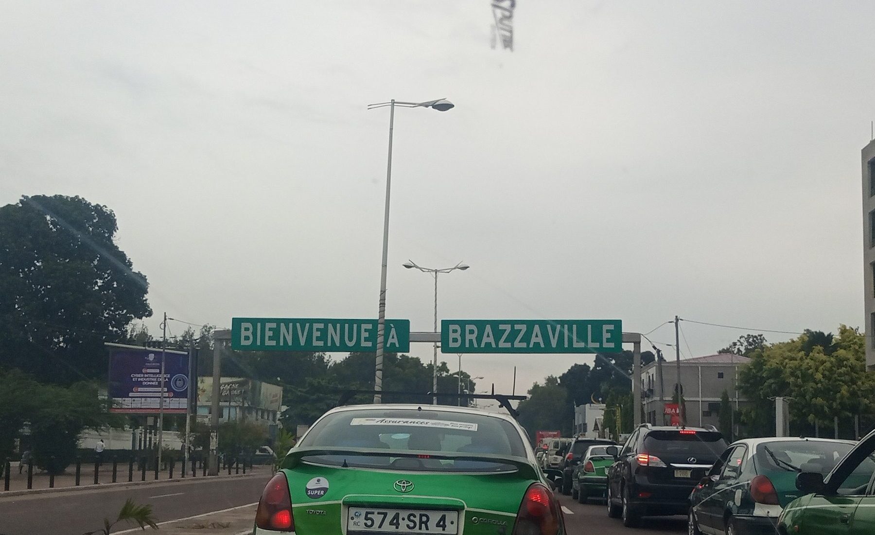 A green taxi in traffic on a Brazzaville road below a sign reading Bienvenue a Brazzaville
