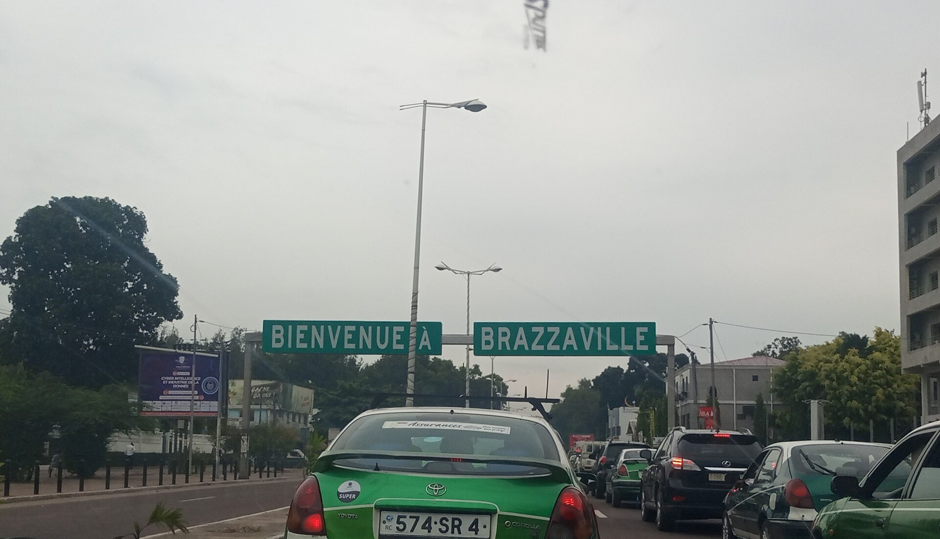 A green taxi in traffic on a Brazzaville road below a sign reading Bienvenue a Brazzaville