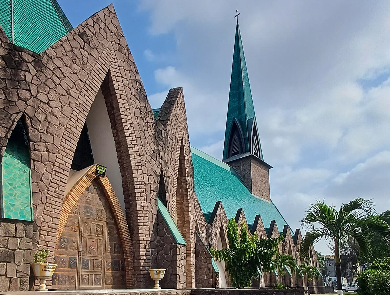 A stone-clad church with a pointed green copper spire and green roof under a partly cloudy sky, palm trees in front