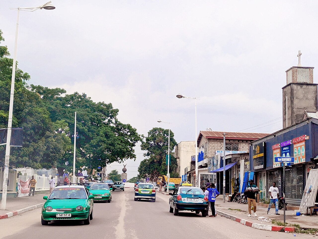 A street in Brazzaville with green and teal taxis, trees on the left and shopfronts and a church tower on the right