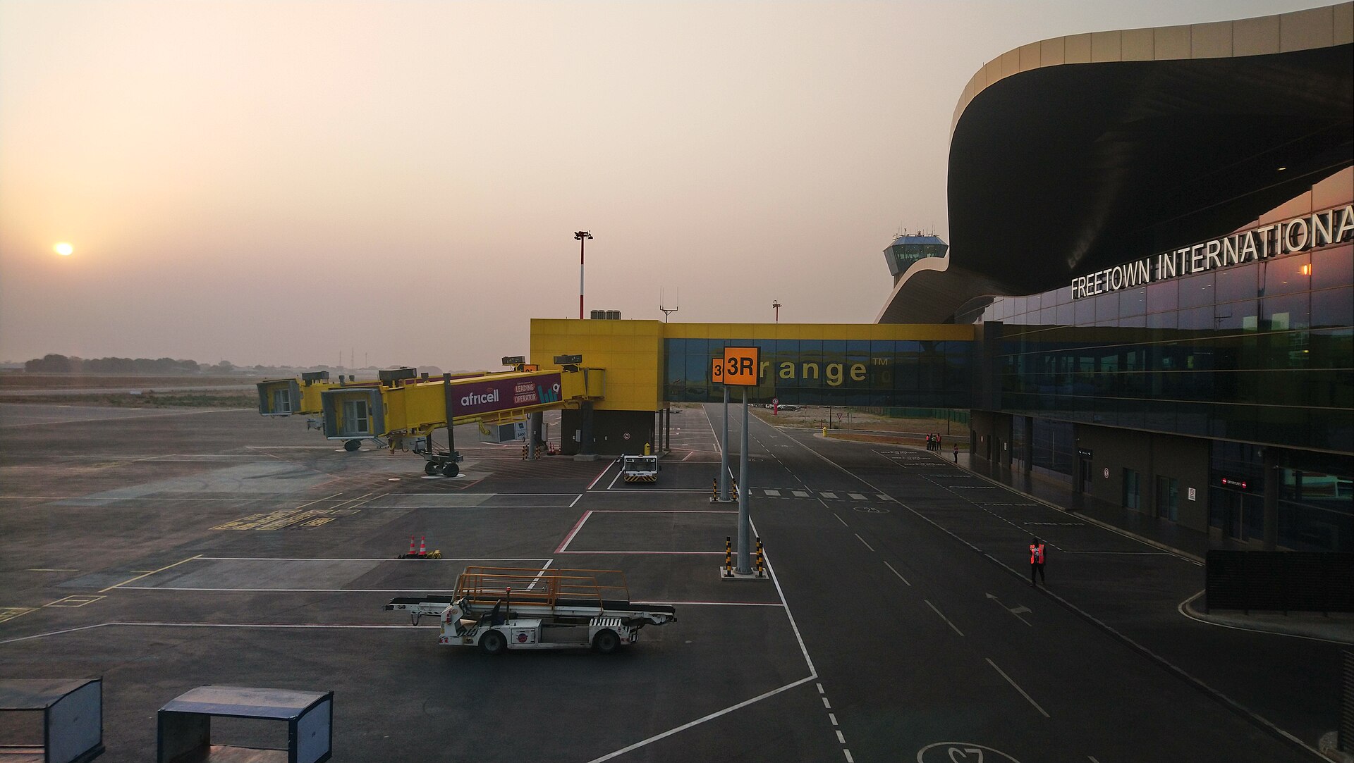 Aircraft stands and passenger boarding bridges beside the glass-fronted Freetown International Airport terminal in hazy low sun
