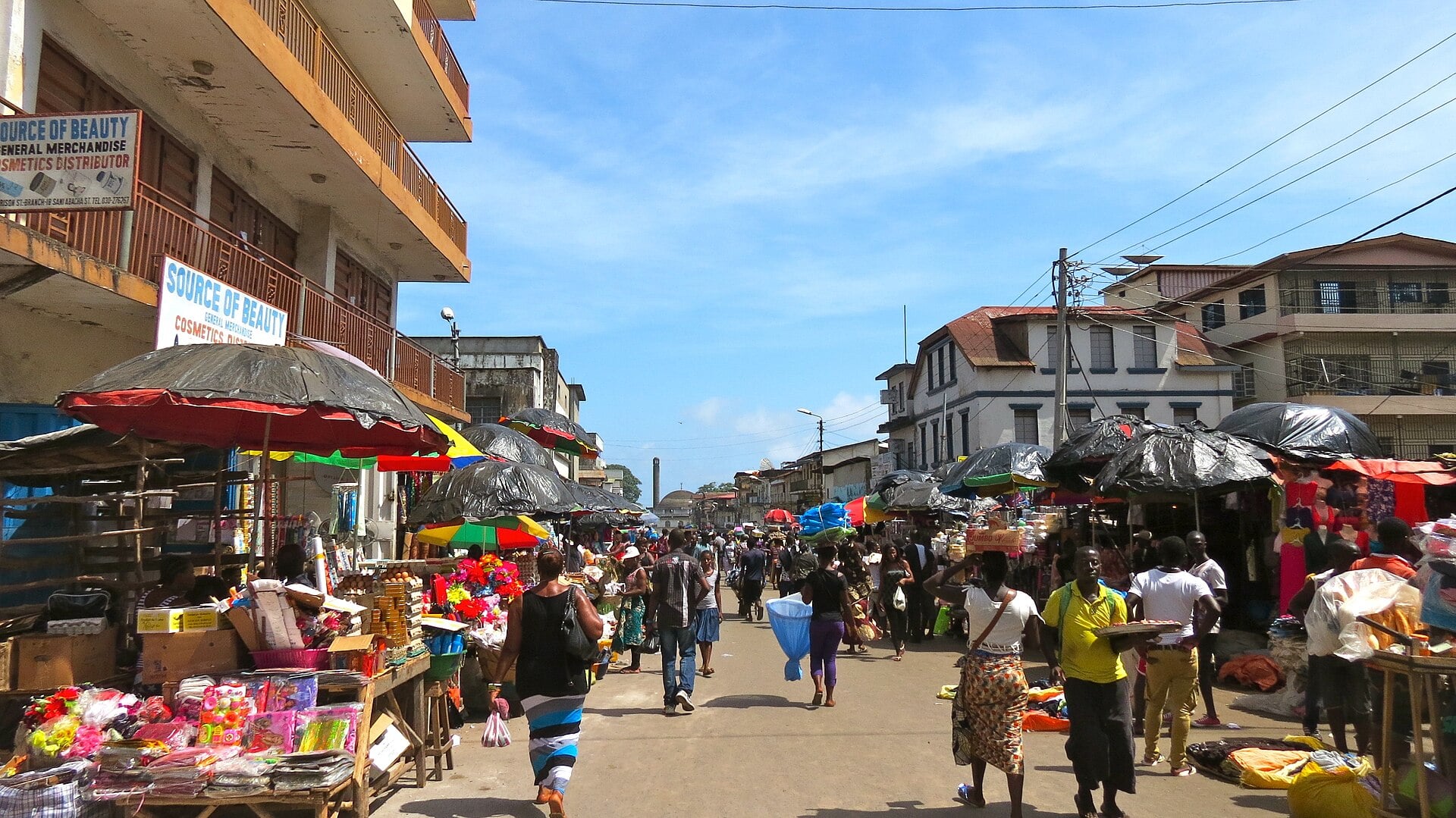 Pedestrians and market stalls with umbrellas along a busy street in central Freetown under a blue sky