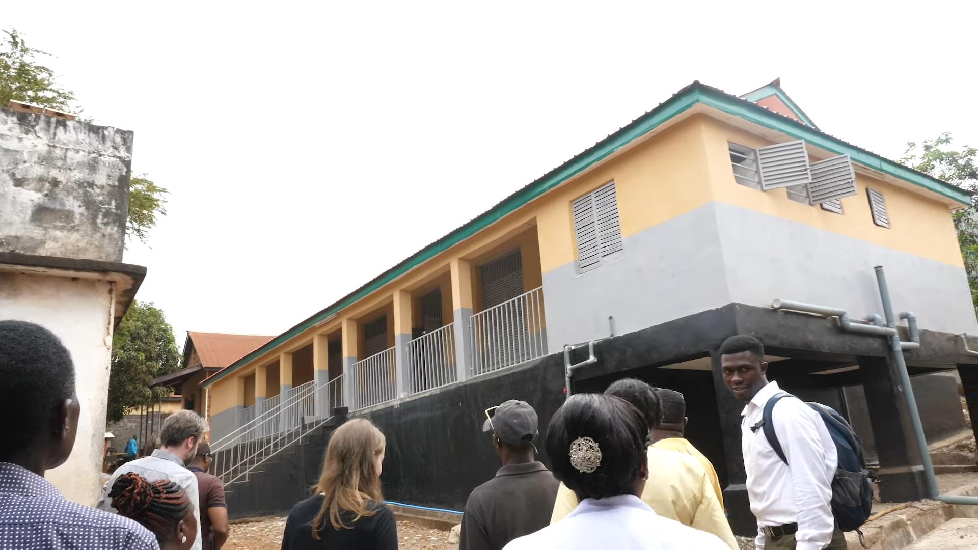 A yellow and grey two-storey hospital building with a green roof and visitors standing in the foreground
