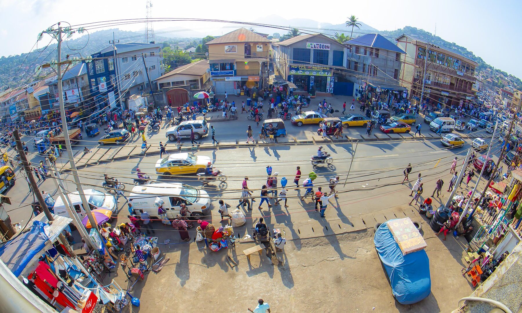 A crowded street in Freetown with taxis, motorbikes, market stalls and green hills behind, seen through a wide-angle lens