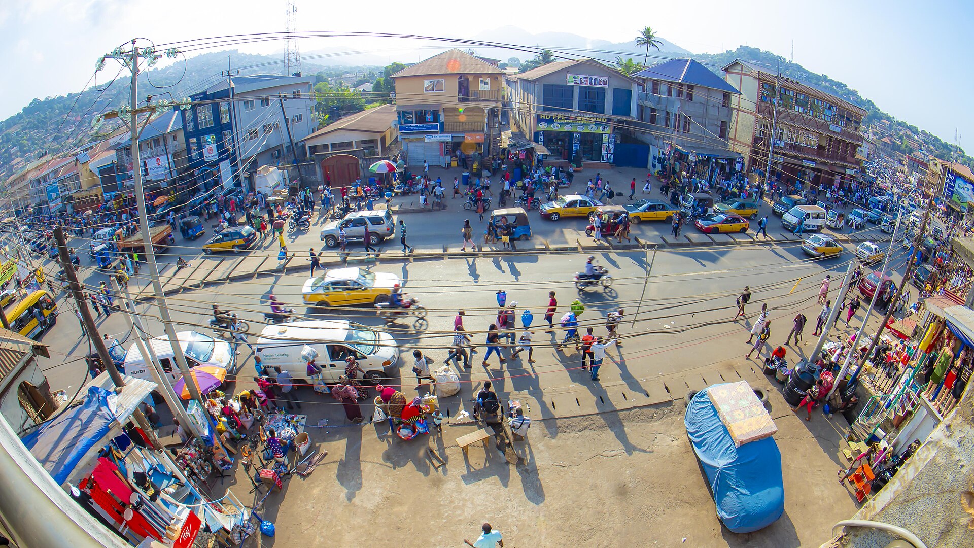 A crowded street in Freetown with taxis, motorbikes, market stalls and green hills behind, seen through a wide-angle lens