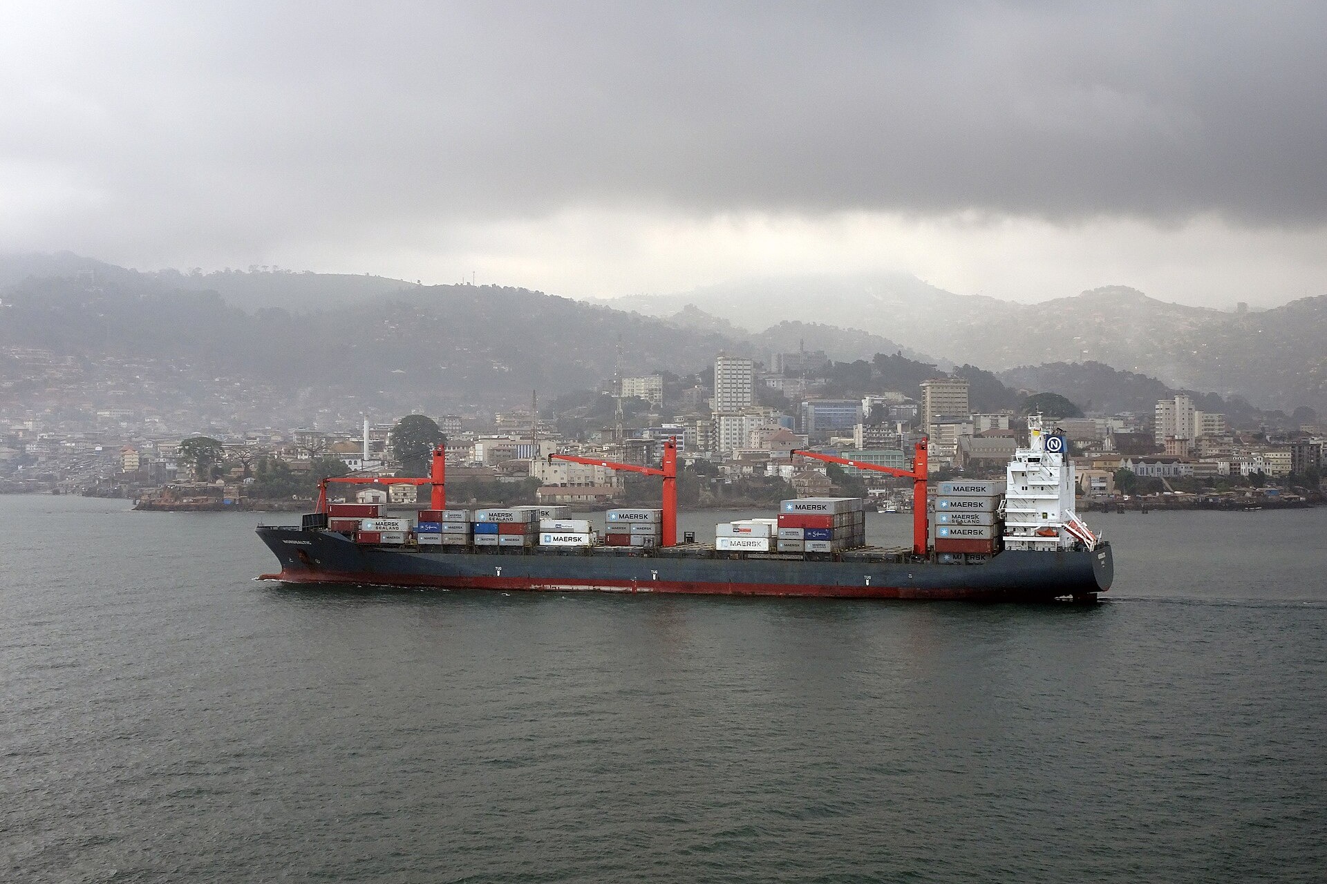 A dark-hulled container ship with red cranes sailing in front of Freetown's hillside buildings under a grey cloudy sky