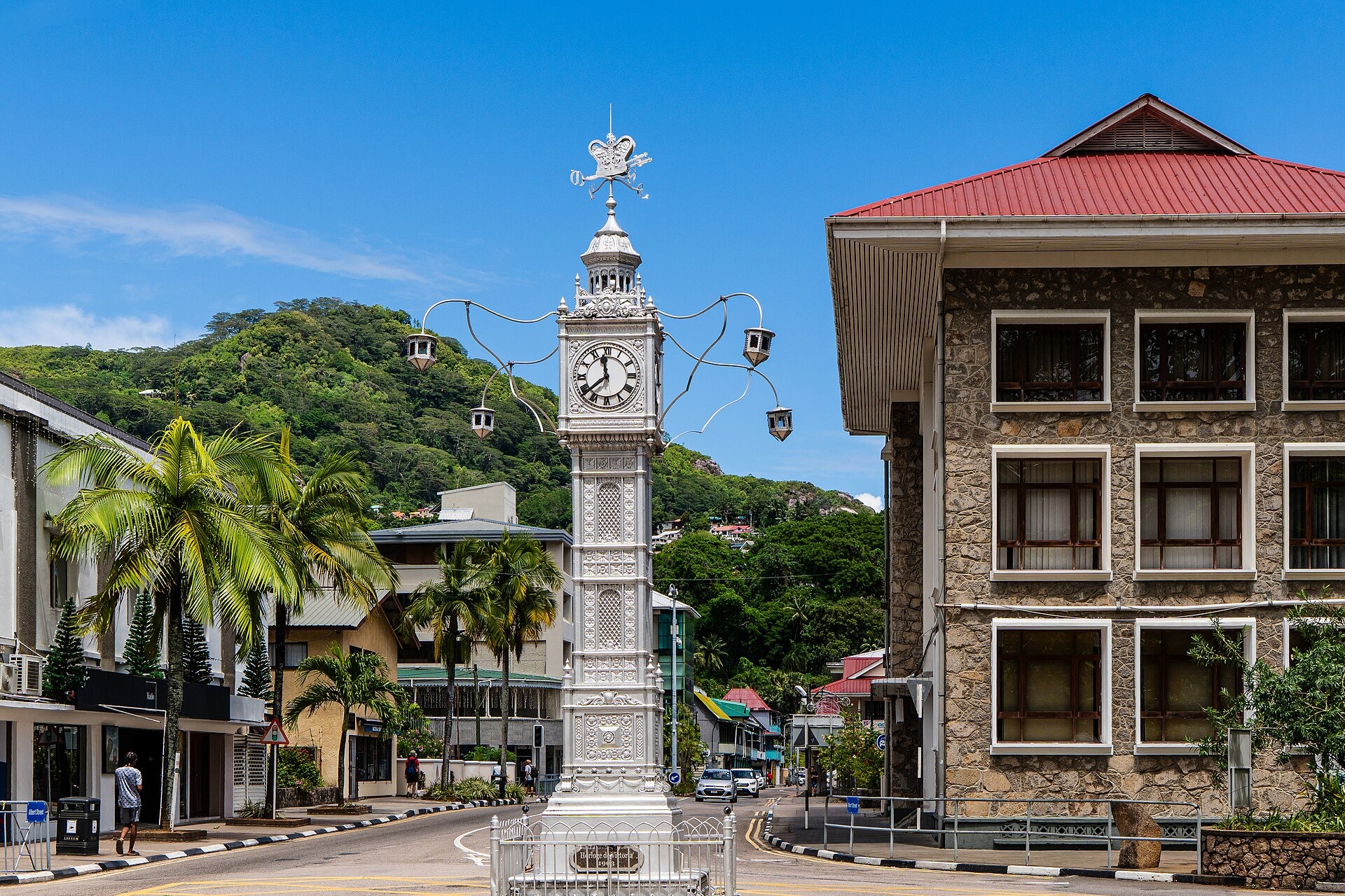 A silver ornamental clock tower on a road junction in Victoria, beside a stone building with a red roof and a green hill behind