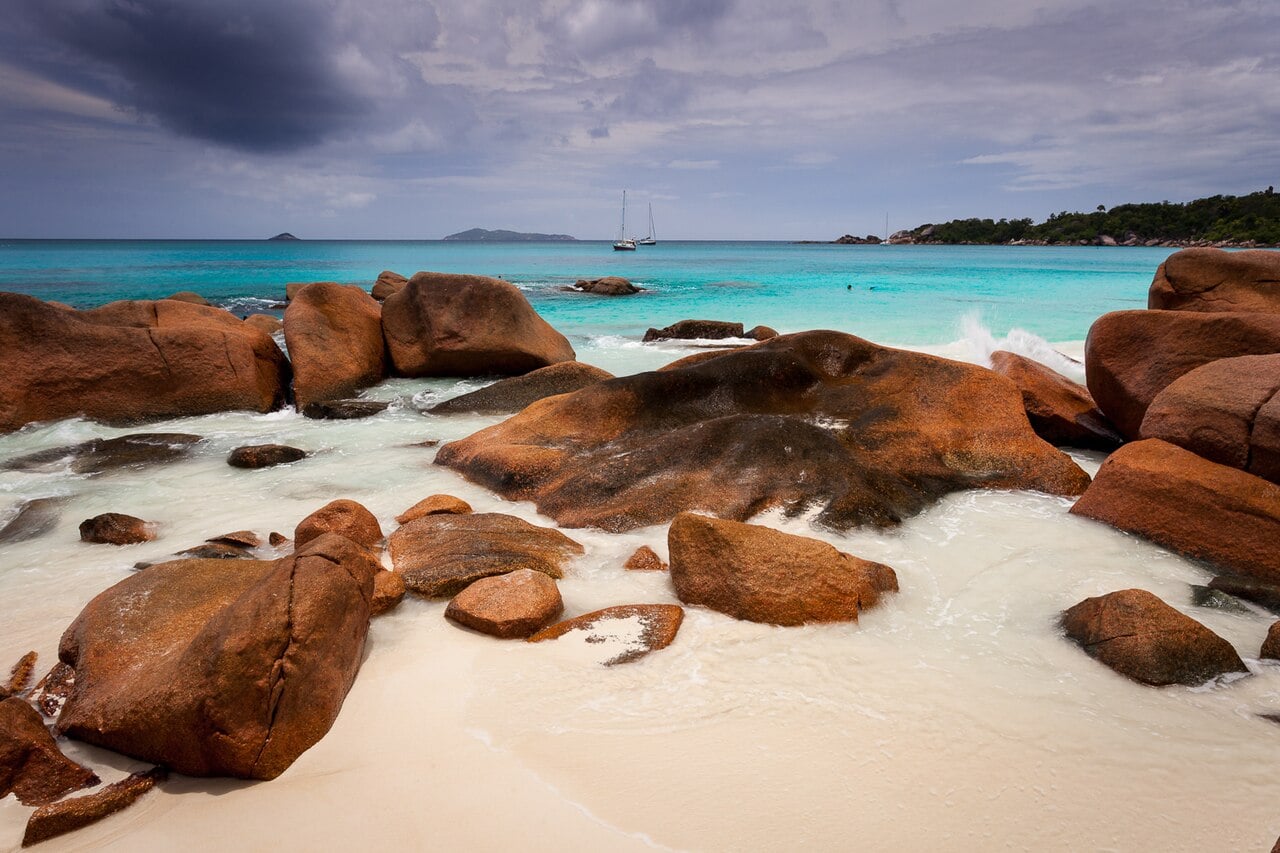 Orange granite boulders on white sand at Anse Lazio beach on Praslin, with turquoise sea and two sailboats beyond