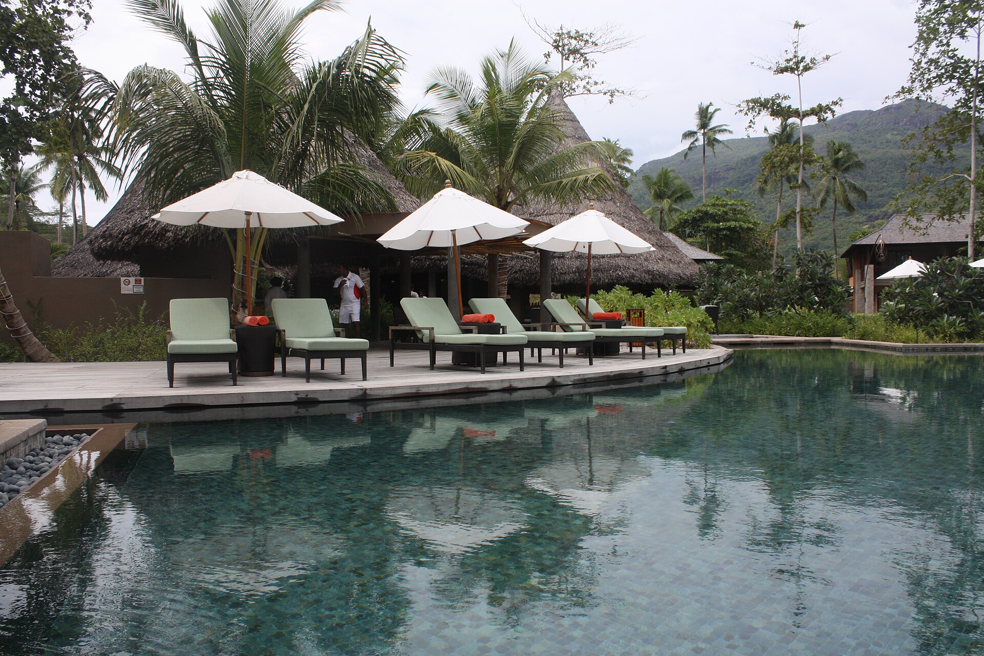 A resort swimming pool with green sun loungers, white parasols and thatched-roof buildings below forested hills