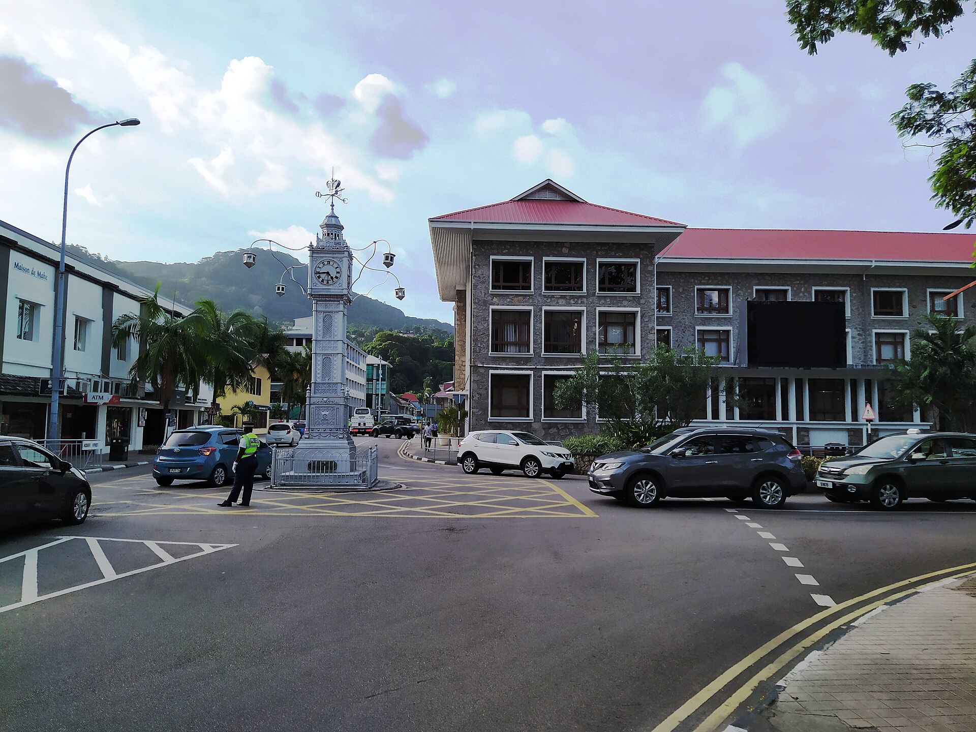 A small silver clock tower on a street junction in Victoria with a red-roofed stone building and a traffic officer