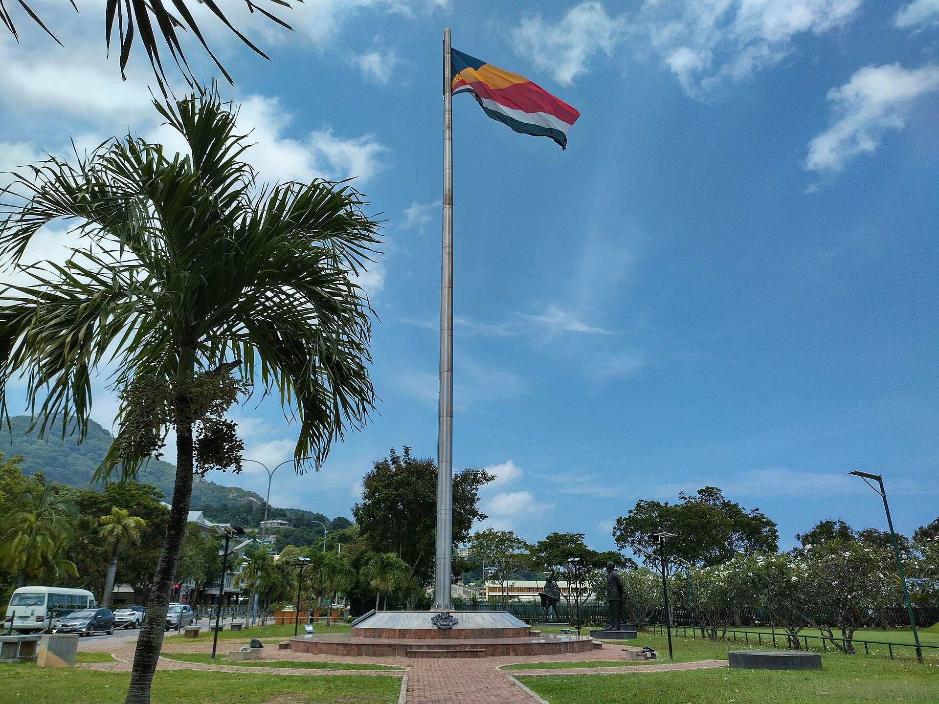 The Seychelles flag flying on a tall pole in a park in Victoria with palm trees, hills and a blue sky