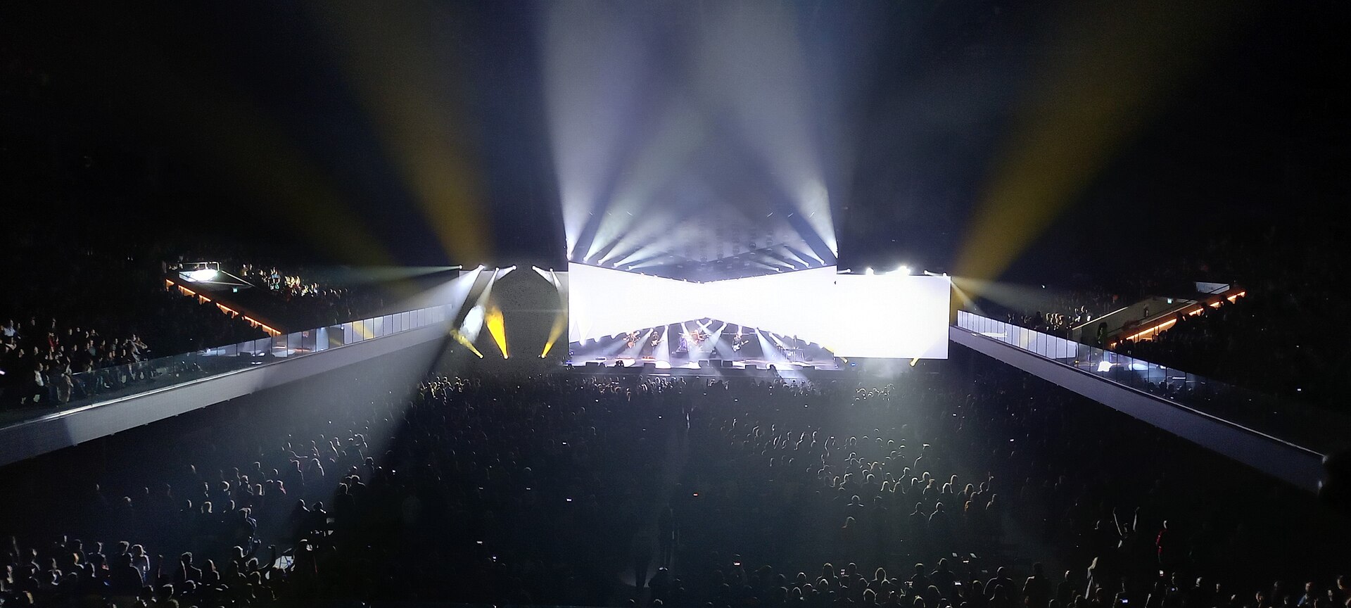 Wide view of a packed arena floor and stands at night, with white and yellow spotlights beaming over a stage with a large lit screen