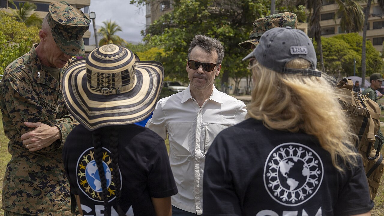 A man in a white shirt and sunglasses talks with women wearing black GEM T-shirts, next to a US Marine general in camouflage uniform