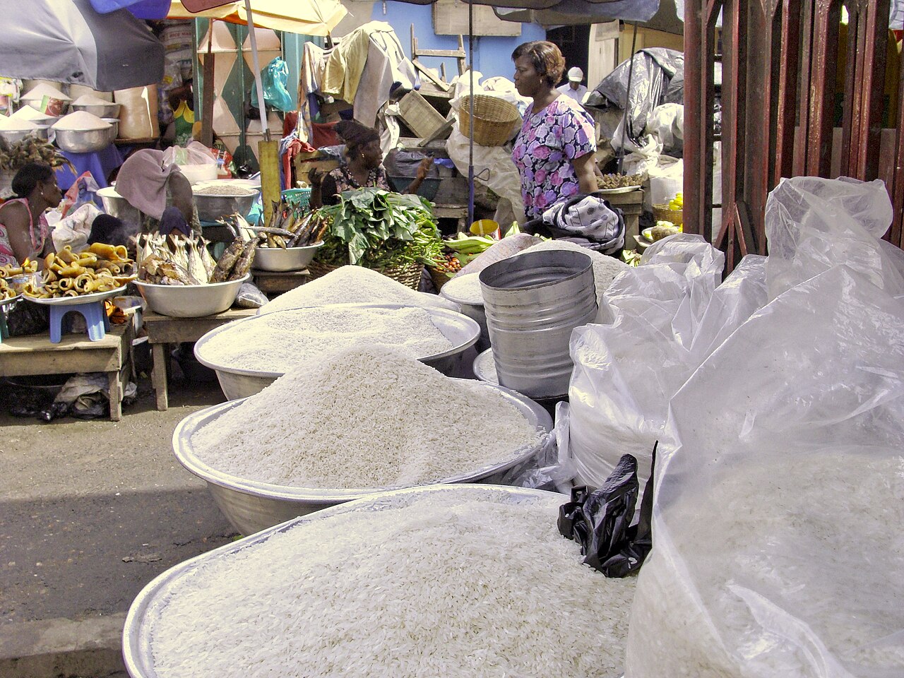Basins heaped with rice, dried fish and vegetables on market stalls in Accra, with a woman standing among the stalls