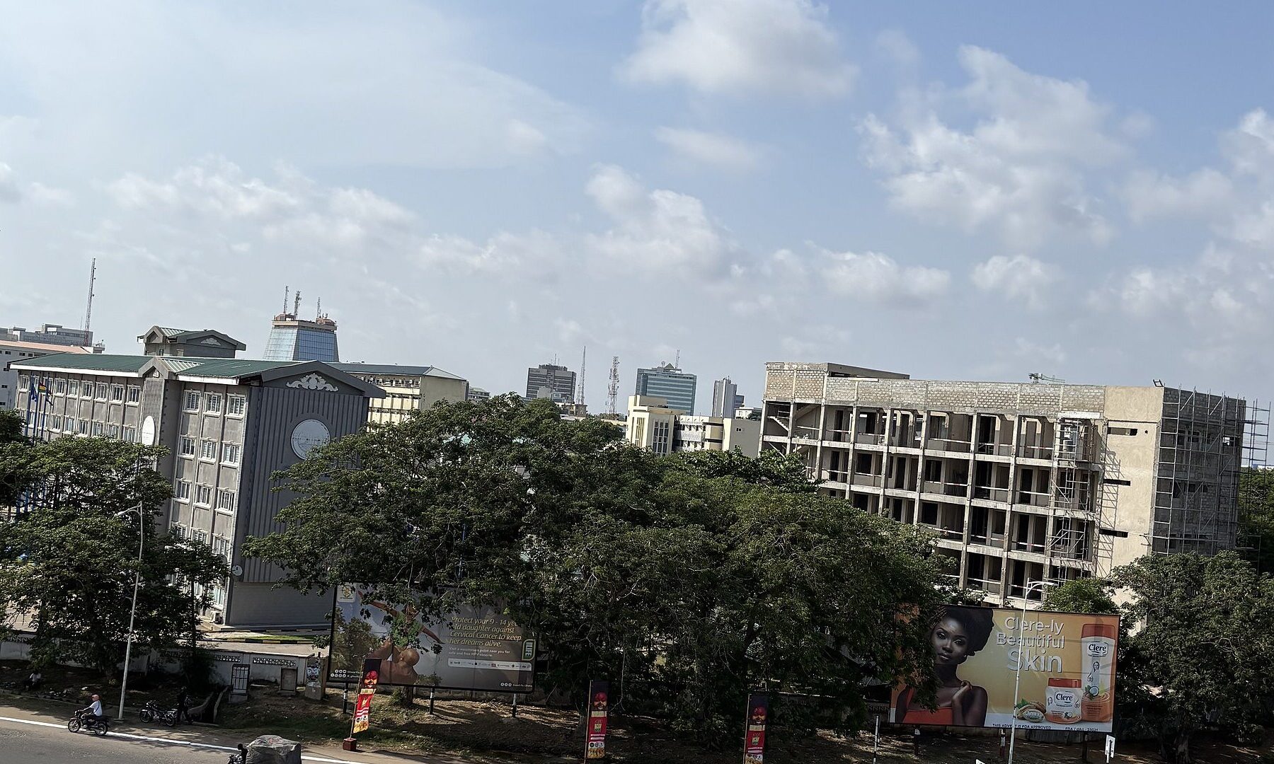 Government and office buildings with trees and an unfinished block under construction in central Accra, Ghana, under a partly cloudy sky